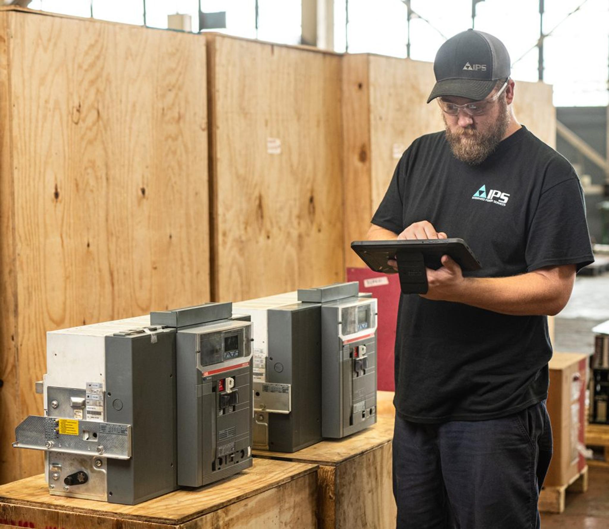 Technicien IPS inspectant des disjoncteurs industriels à l’aide d’une tablette dans un atelier.