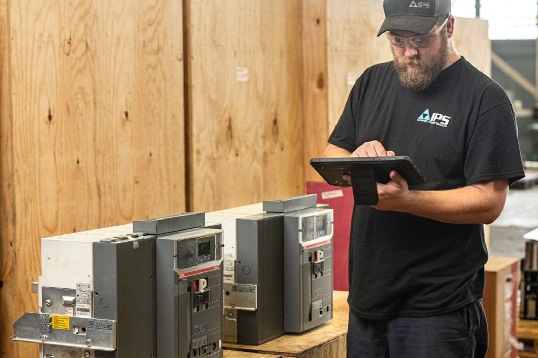 IPS technician uses a tablet while inspecting industrial circuit breakers in a service facility.