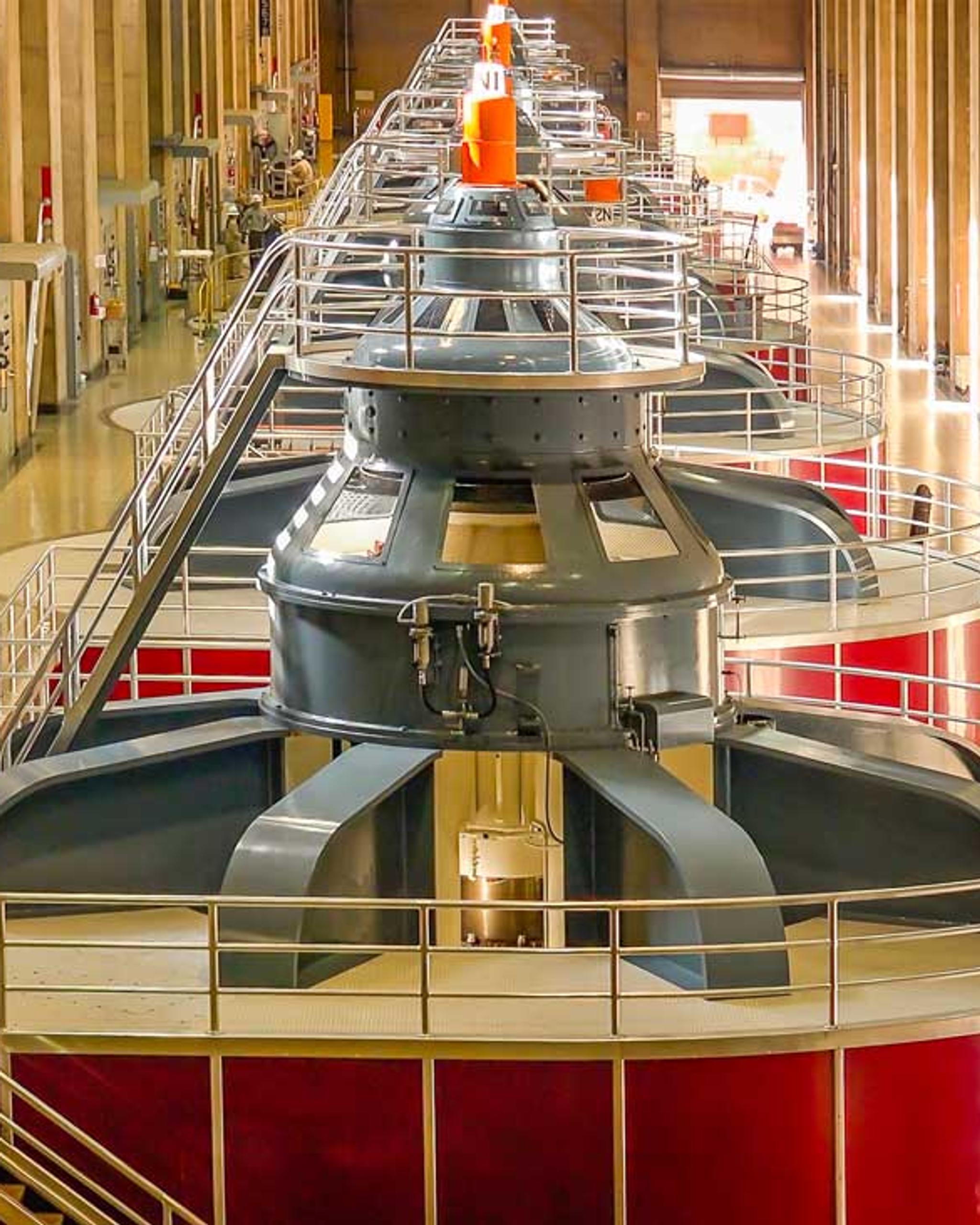 Fila de grandes generadores hidroeléctricos con barandillas y escaleras en una central eléctrica.
