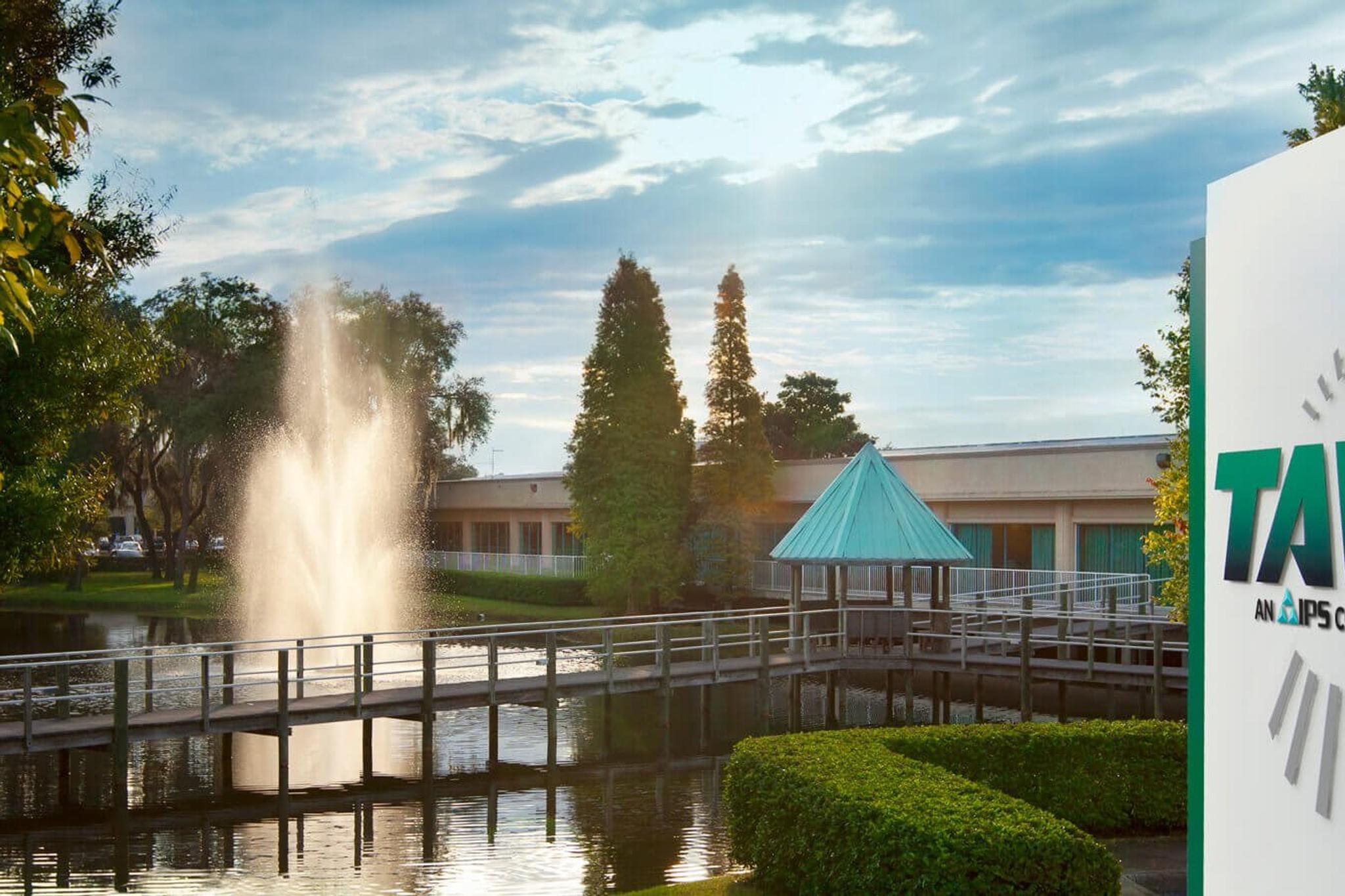 TAW, an IPS company, facility sign beside a landscaped pond with a fountain and footbridge.