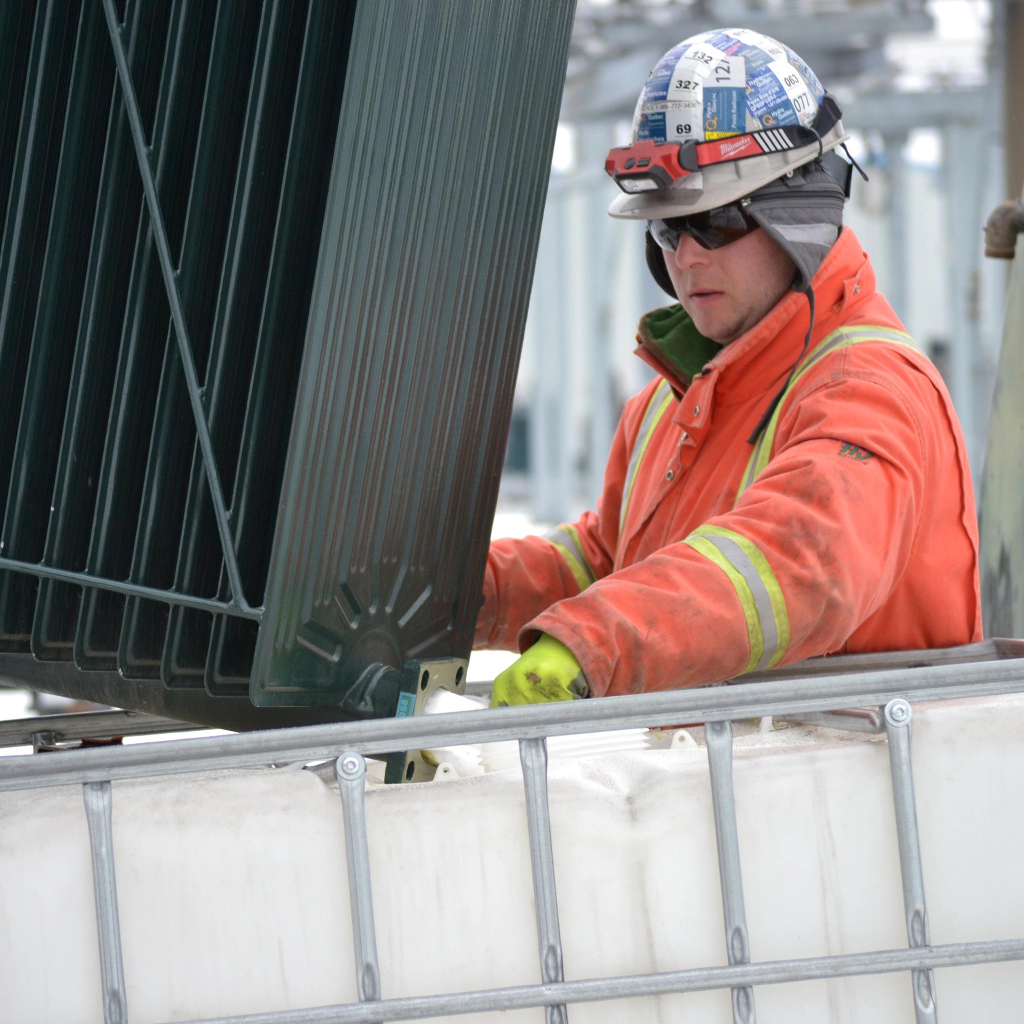 Technician in safety gear services a large transformer radiator beside an industrial fluid tank.