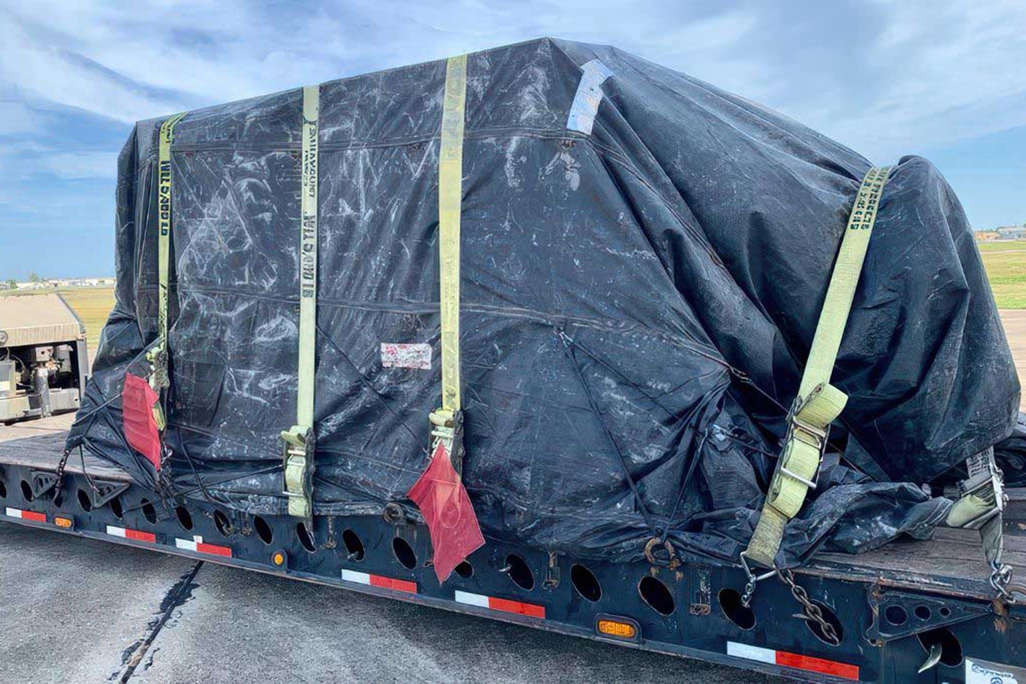 Large industrial equipment covered by a black tarp and secured with straps on a flatbed trailer.
