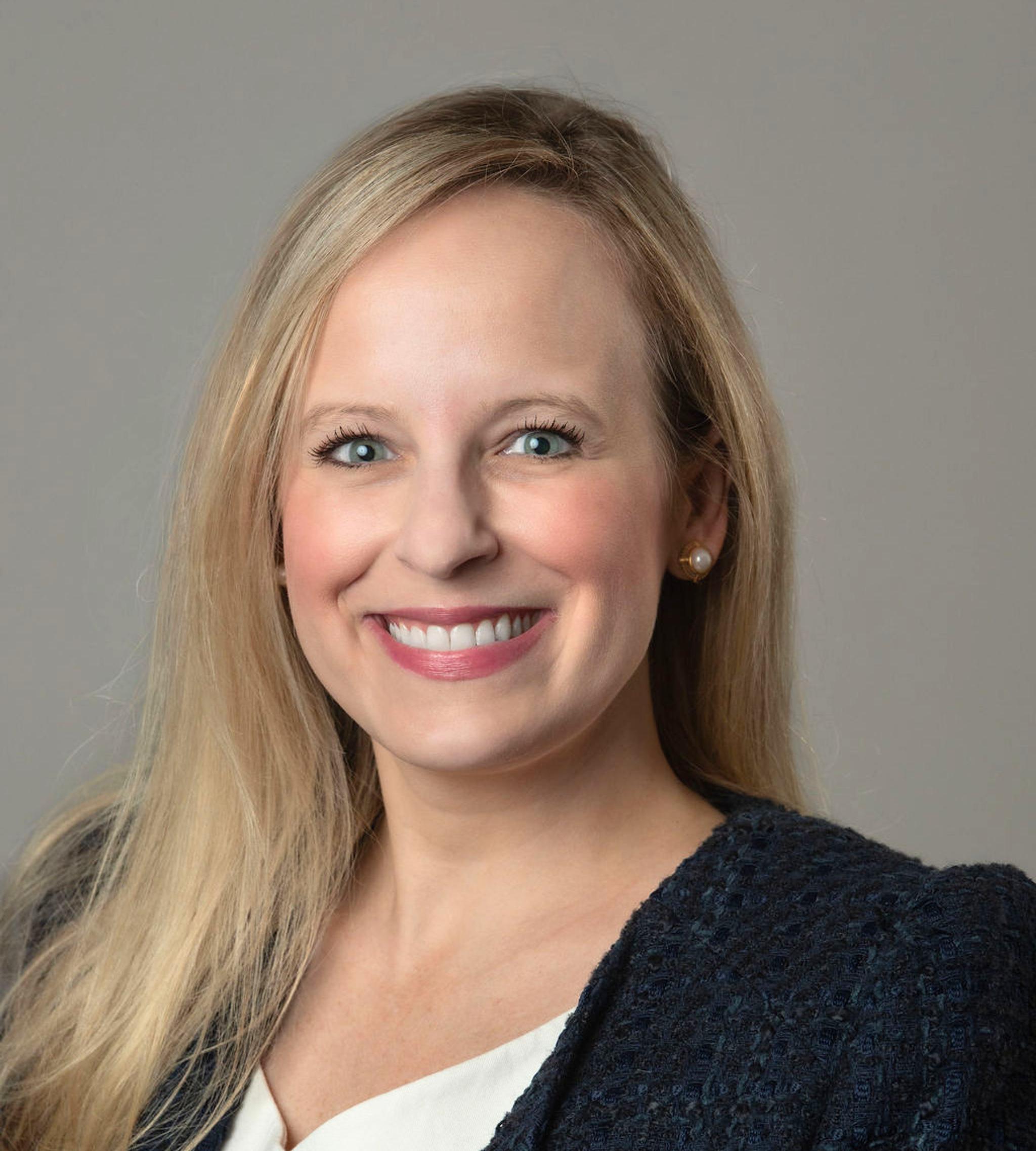 Professional headshot of a smiling blonde woman in a navy blazer and white blouse.