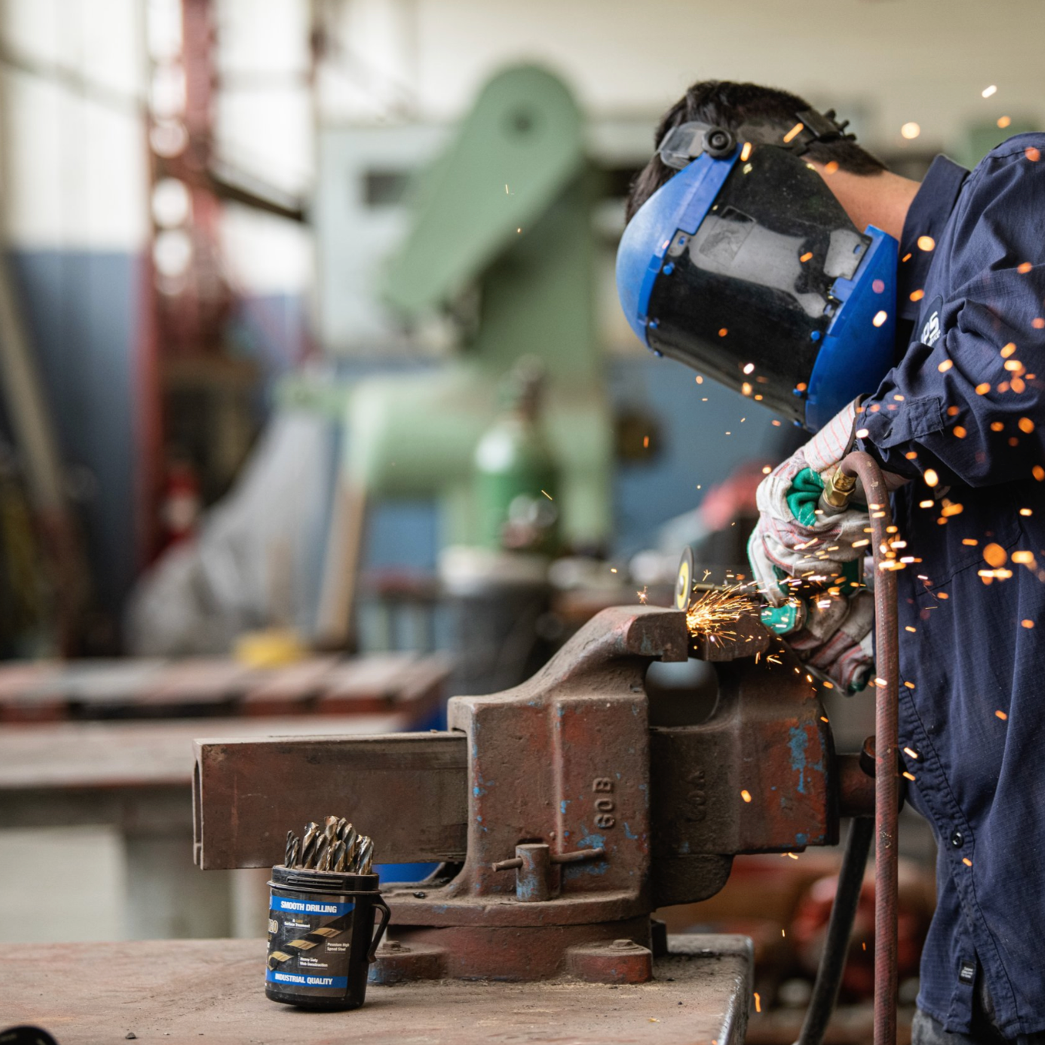 Technician wearing a face shield grinds metal held in a bench vise as sparks fly.