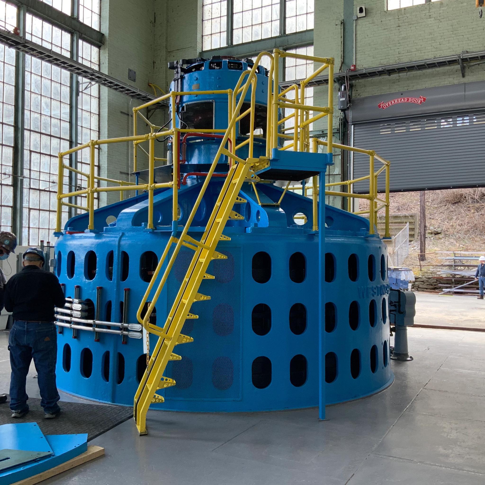 Technicians inspect a large blue generator with yellow access stairs inside an industrial facility.