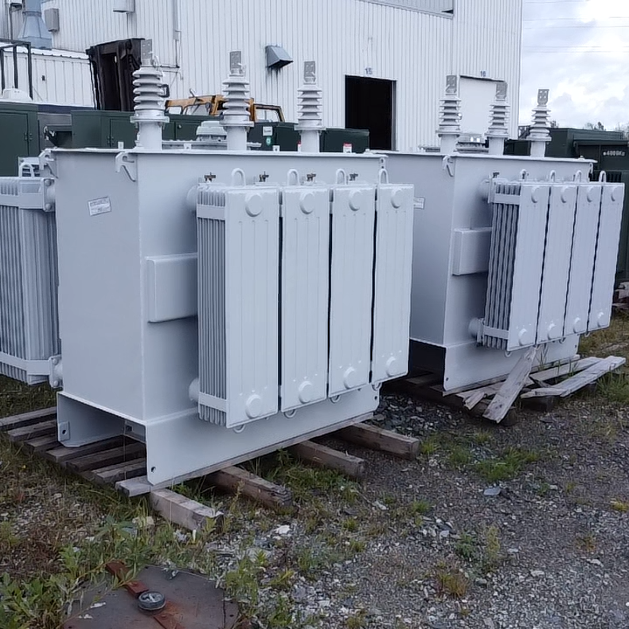 Two gray power transformers with cooling radiators and bushings stand outside an industrial service facility.