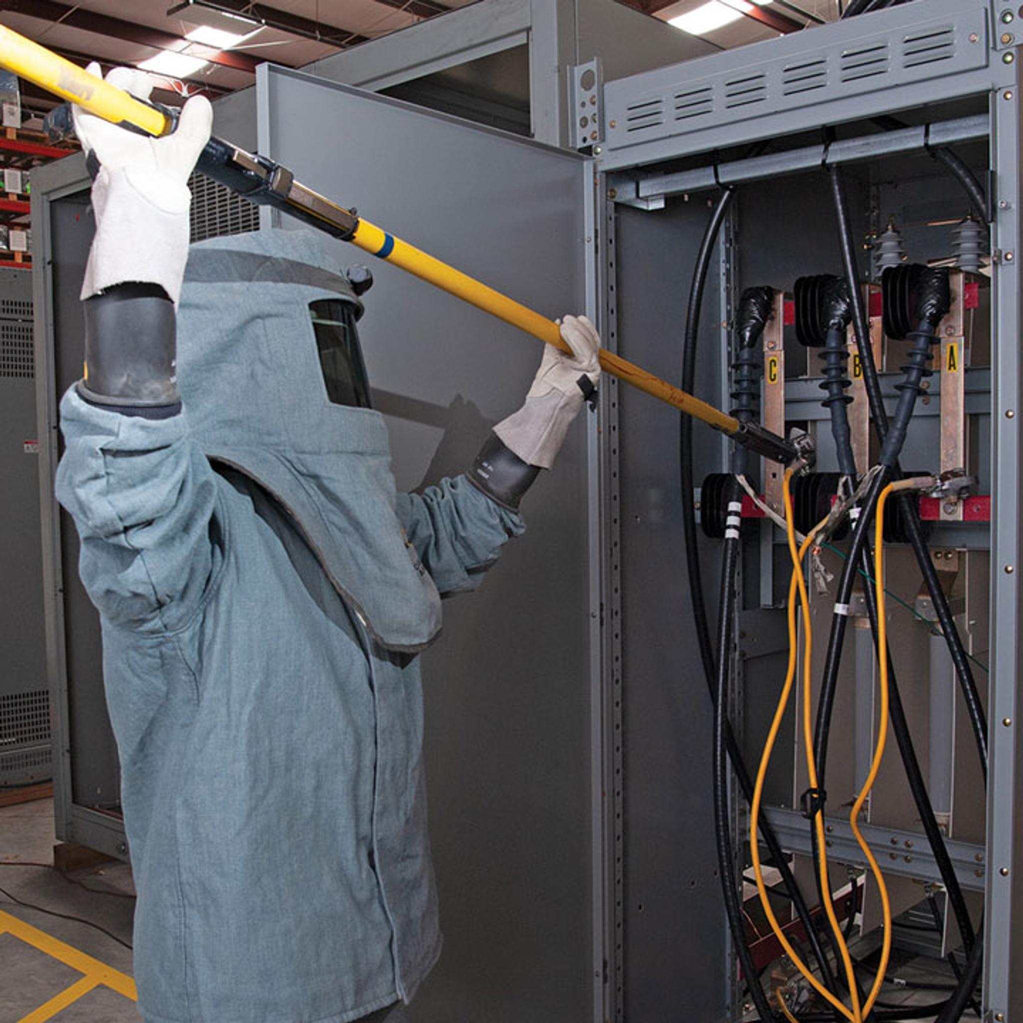 Technician in arc-flash PPE uses an insulated hot stick to service open switchgear.