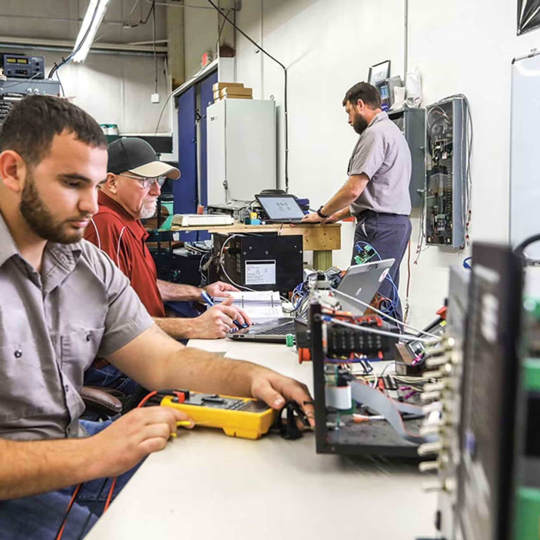 Technicians test and troubleshoot electrical control equipment with multimeters and laptops.
