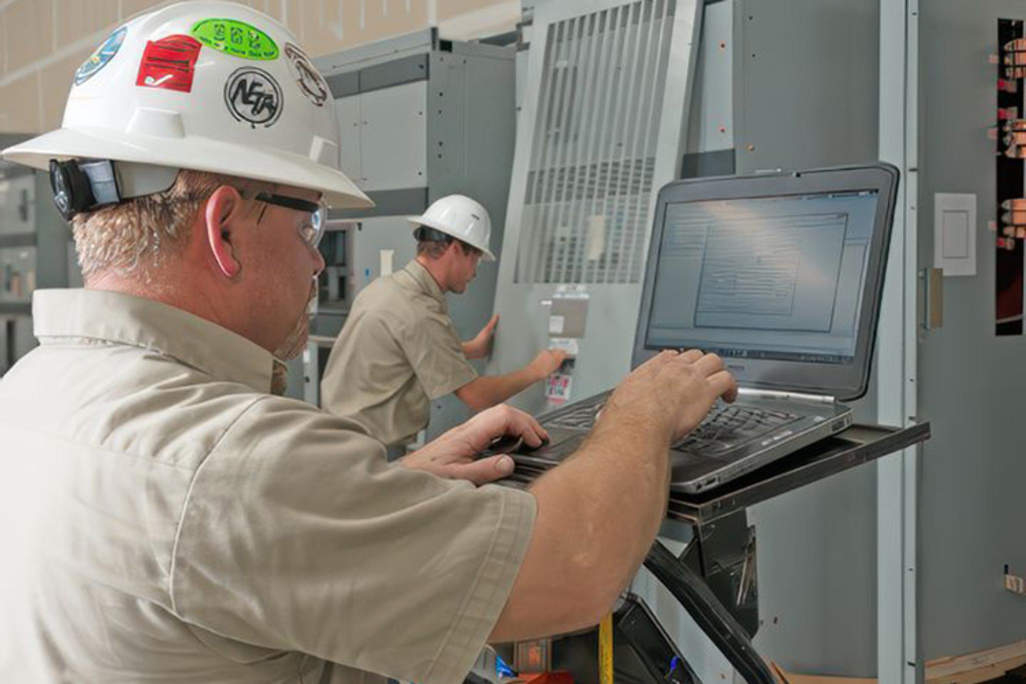Technicians use a laptop to inspect and service industrial electrical equipment.