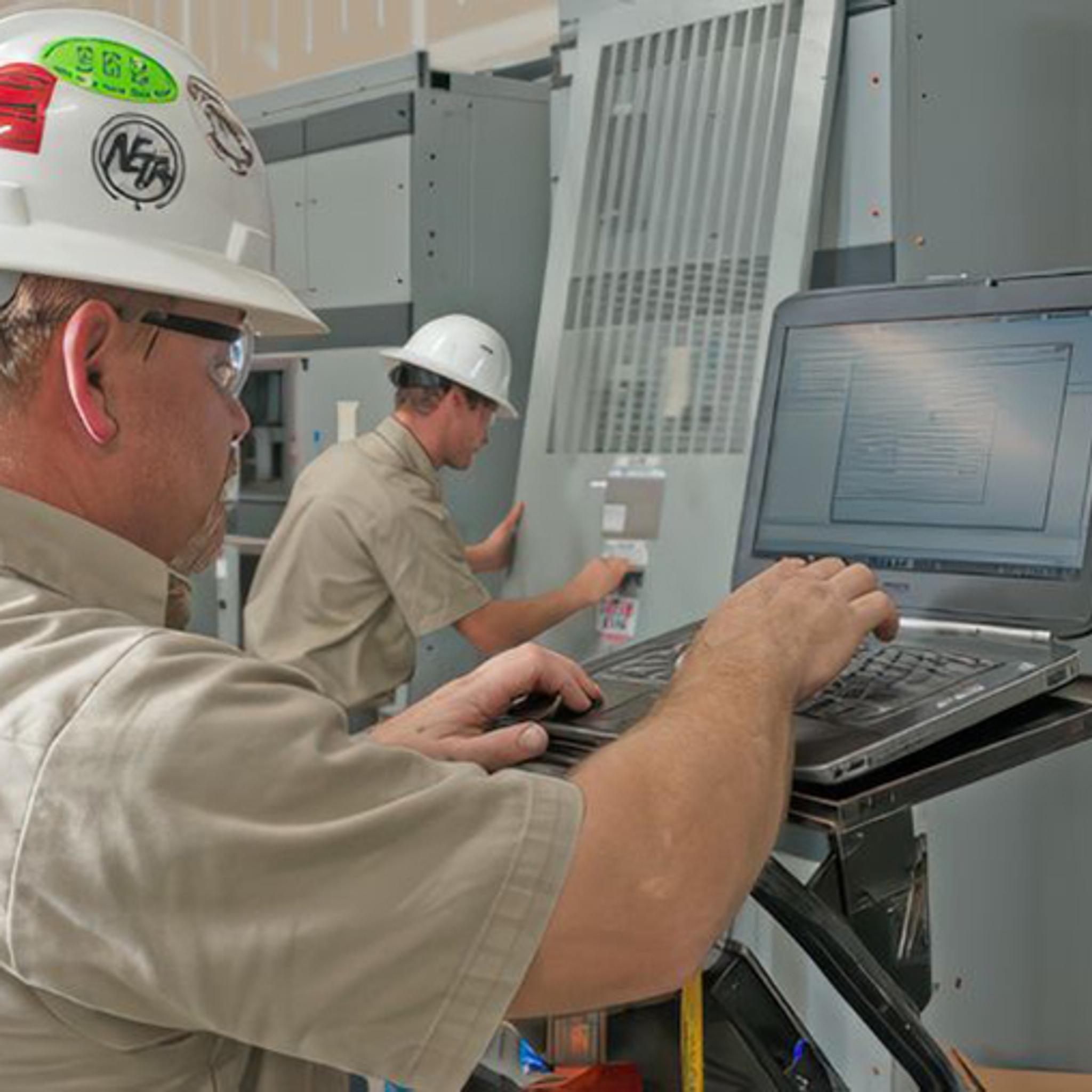 Technicians use a laptop to inspect and service industrial electrical equipment.