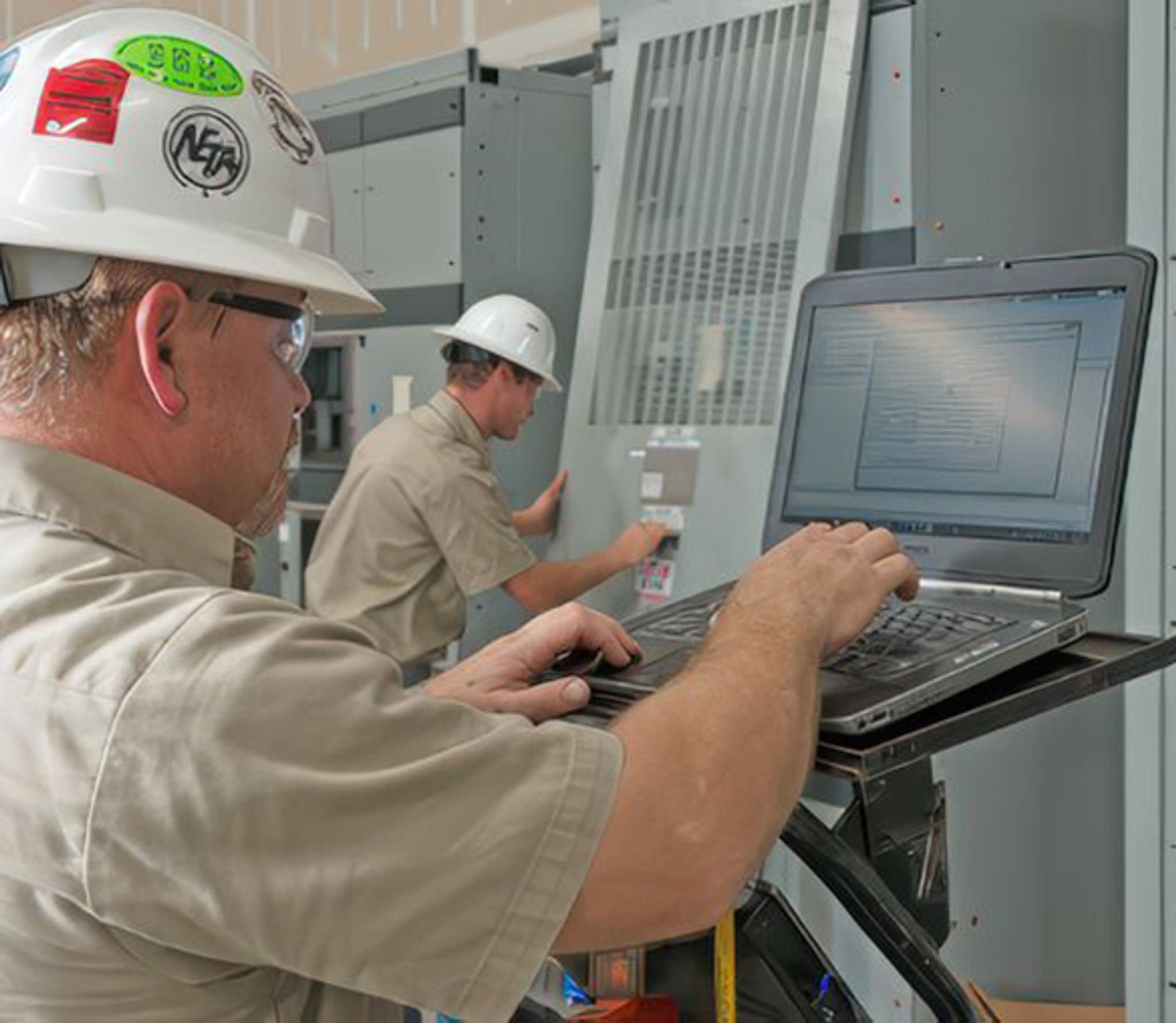 Deux techniciens casqués diagnostiquent une armoire électrique industrielle à l’aide d’un ordinateur portable.