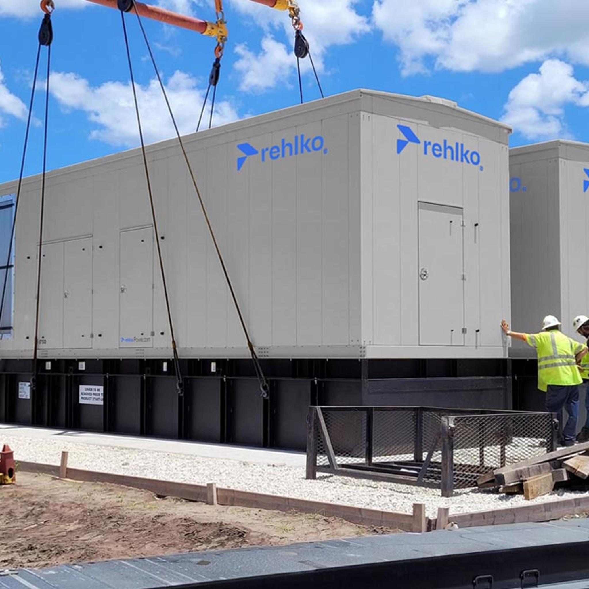 Workers guide a crane as it installs large Rehlko generator enclosures at an industrial site.