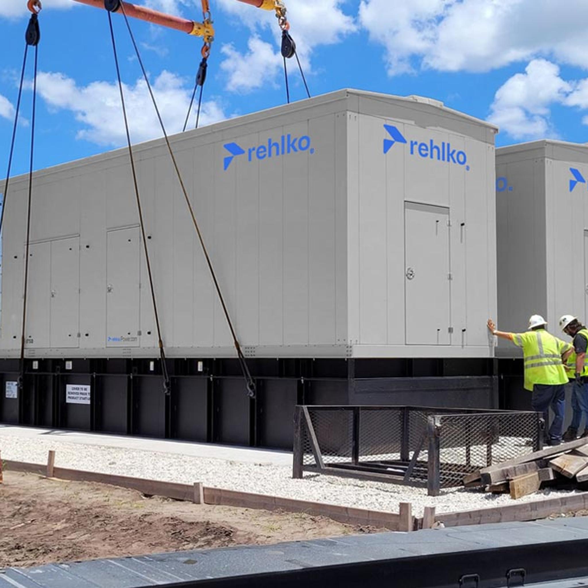 Workers guide a crane as it installs large Rehlko generator enclosures at an industrial site.