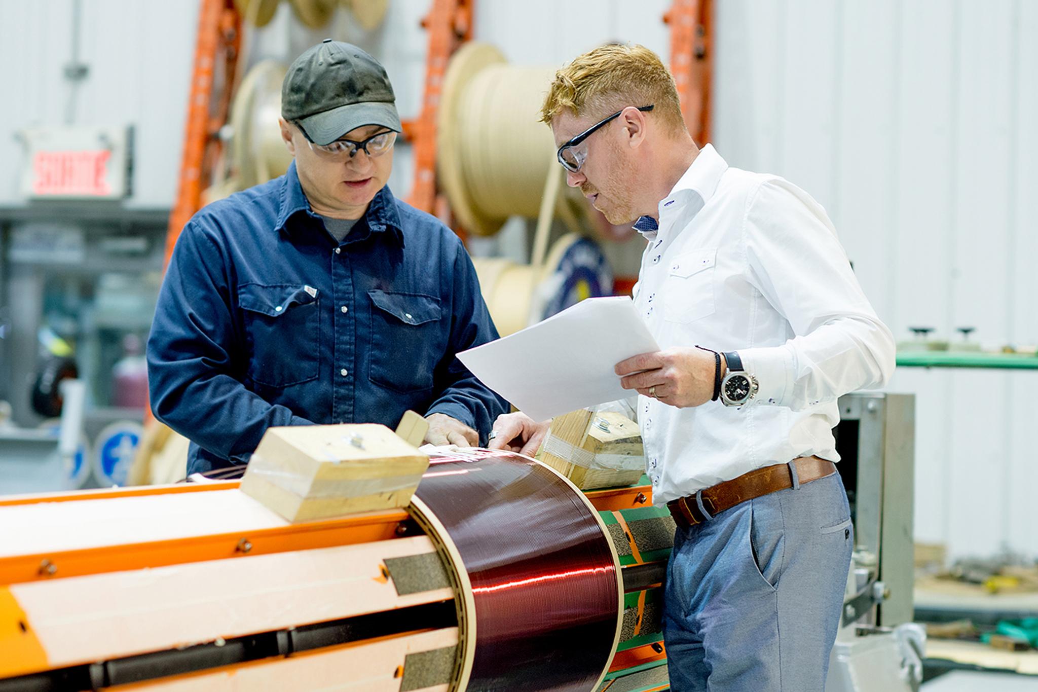 Two technicians inspect copper windings and review documents on a large electric motor stator.
