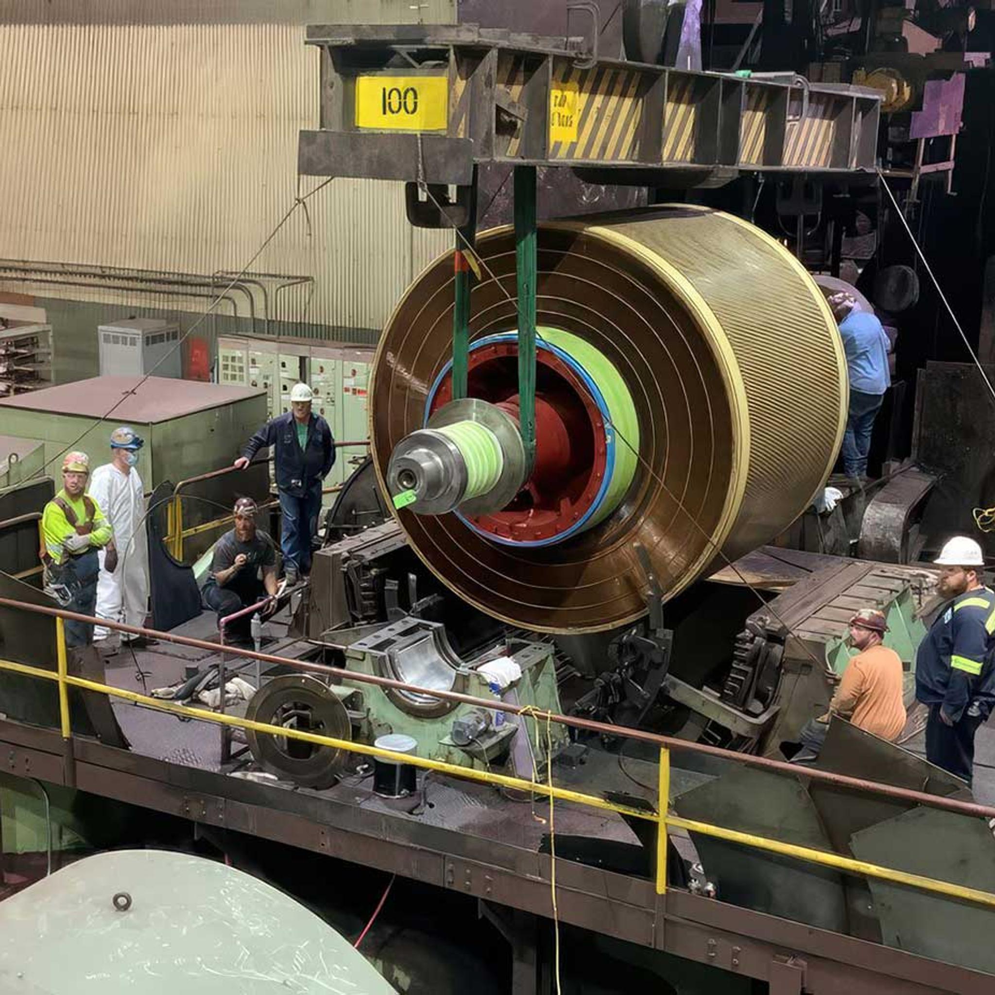 Technicians install a large generator rotor using an overhead crane in an industrial service facility.