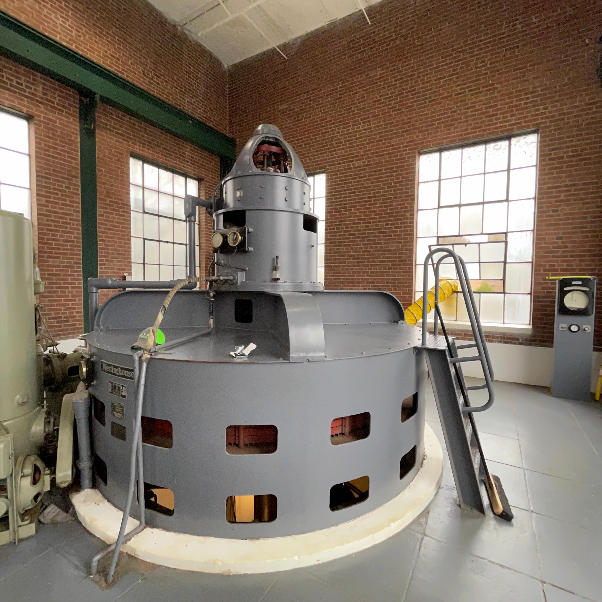 Large Westinghouse generator with gauges and access stairs inside a brick-walled power station.