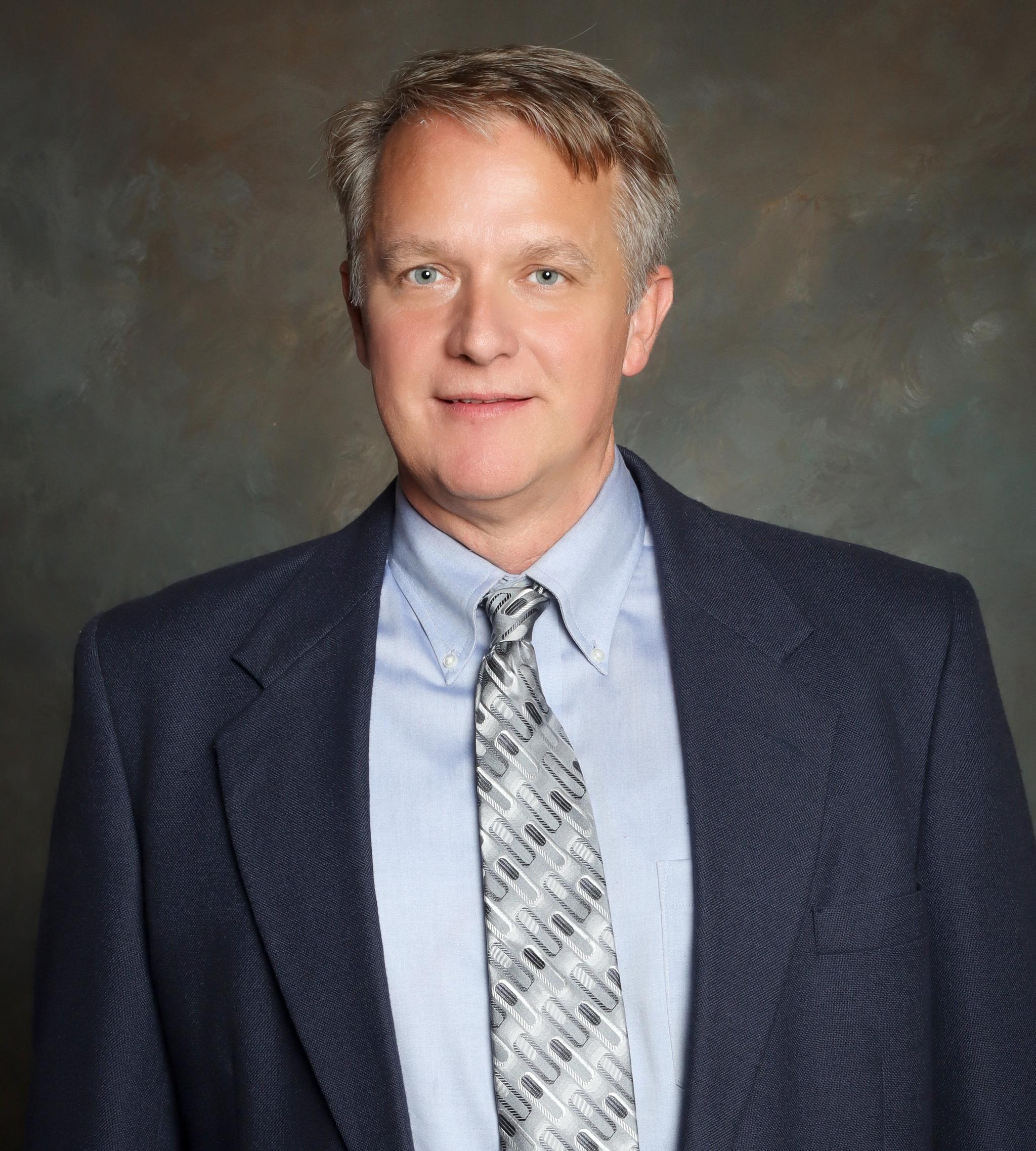 Professional headshot of a man wearing a navy suit, light blue shirt, and patterned tie.