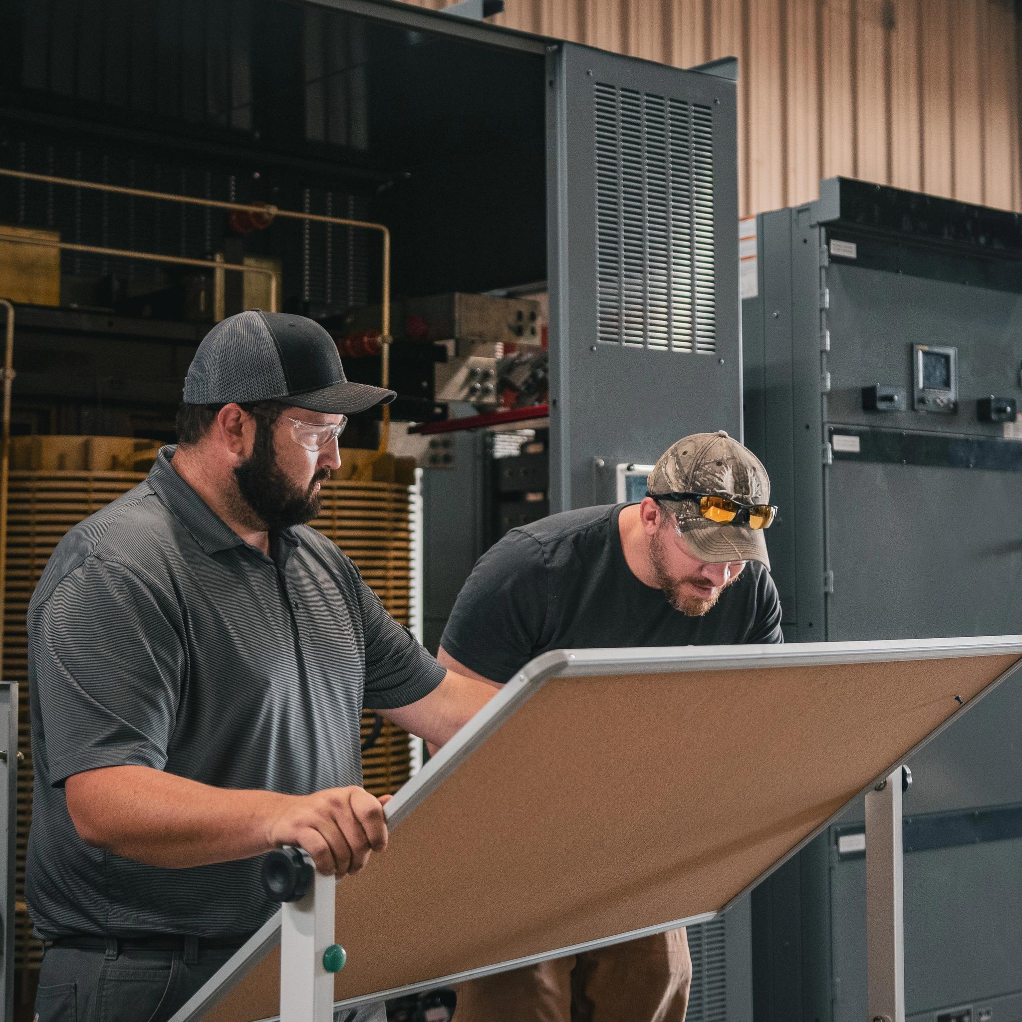 Two technicians review plans beside industrial switchgear cabinets in an electrical service facility.