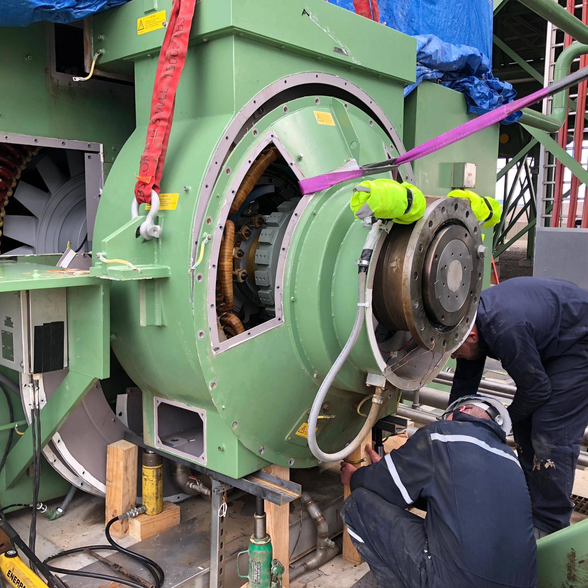 Technicians service a large industrial generator secured with lifting straps and hydraulic jacks.