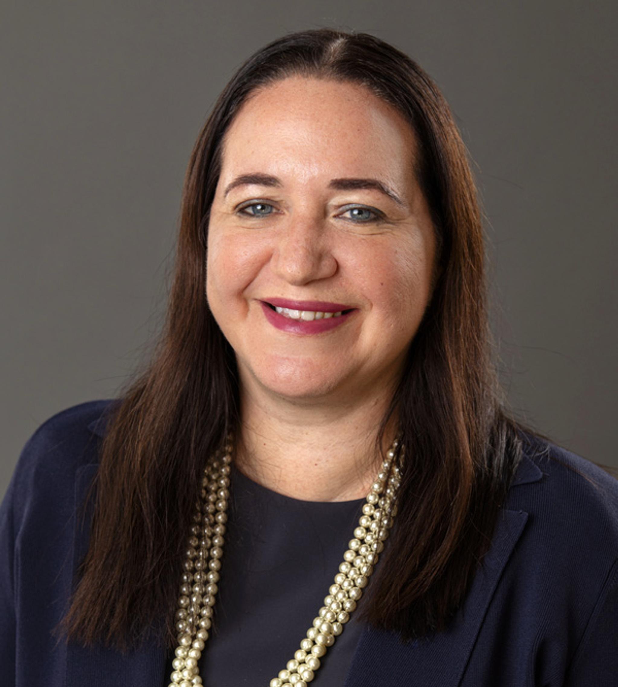Professional headshot of a smiling woman in a navy blazer and pearl necklace.
