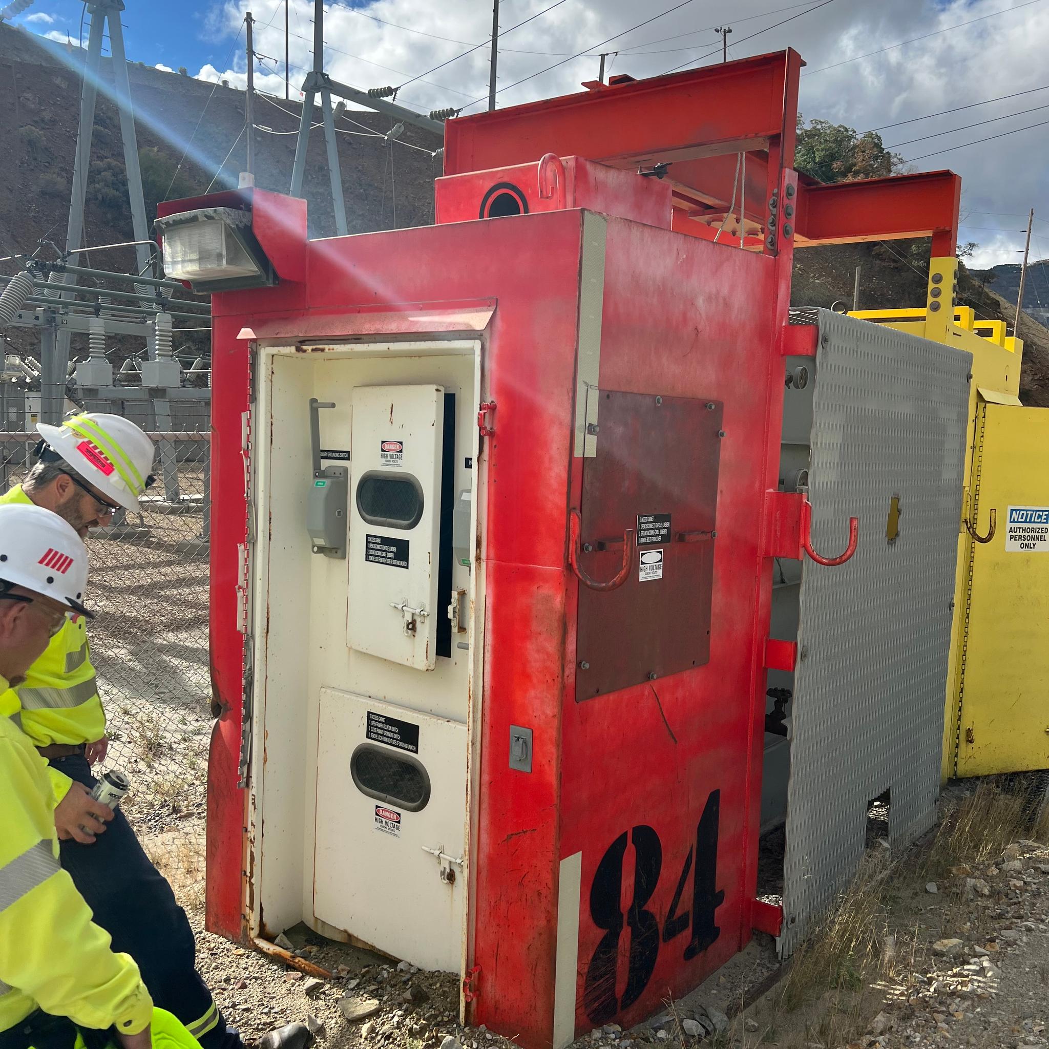 Technicians in safety gear inspect red medium-voltage switchgear at an outdoor electrical substation.
