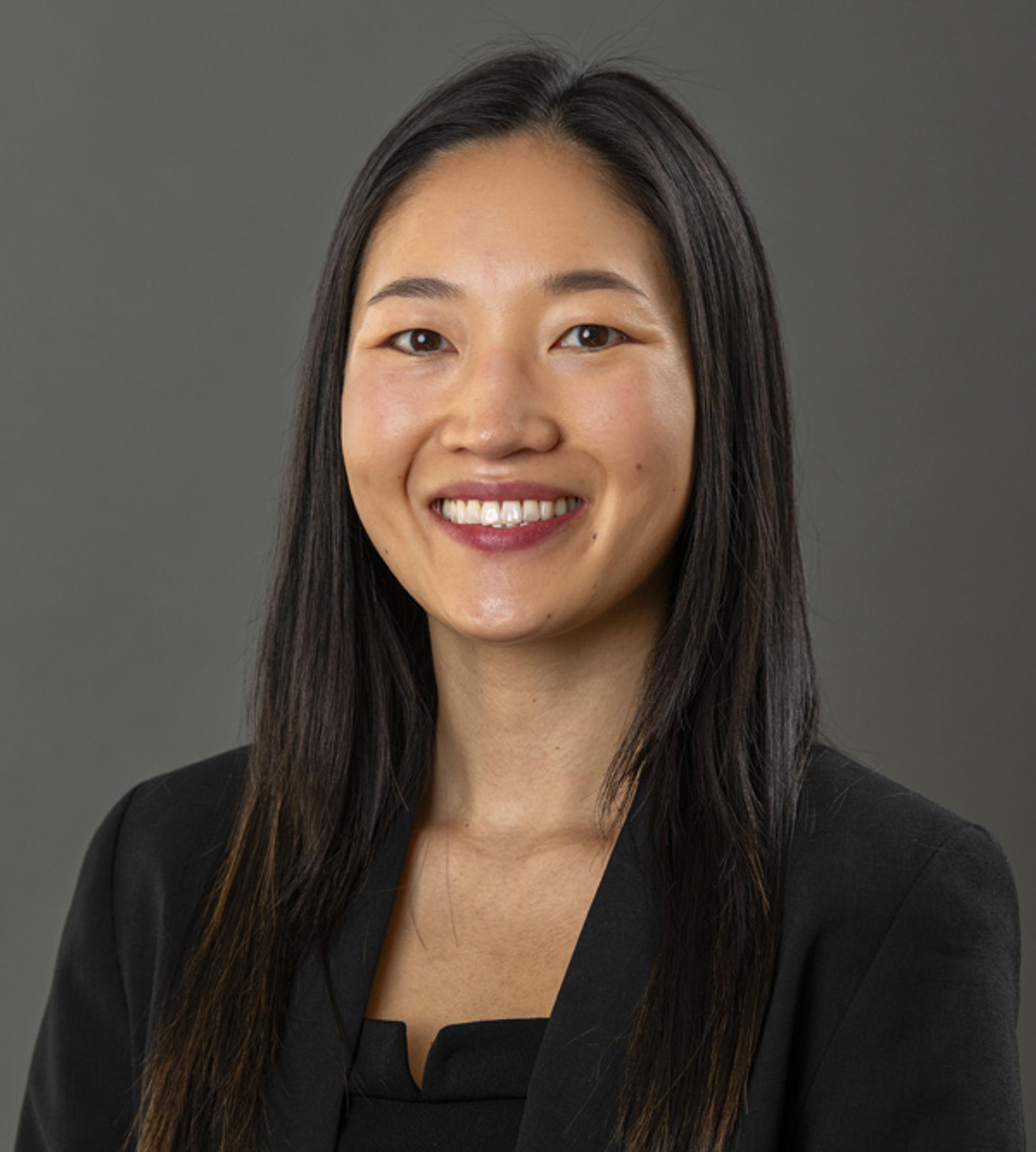 Professional headshot of a smiling woman with long dark hair wearing a black blazer.