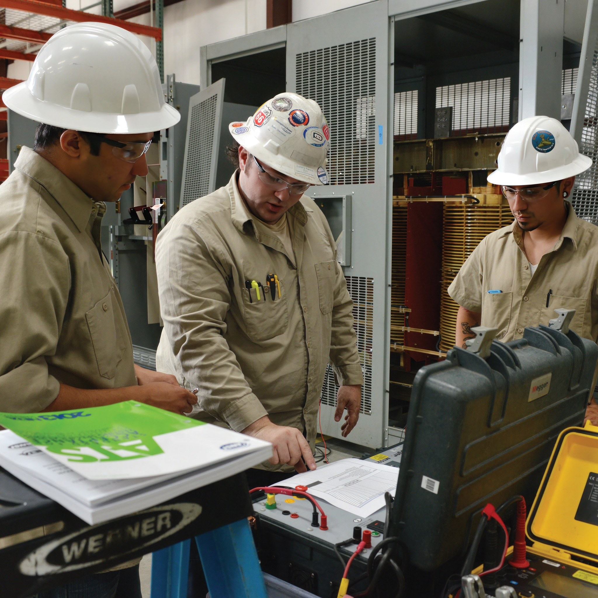 Technicians review electrical test equipment beside open industrial switchgear.
