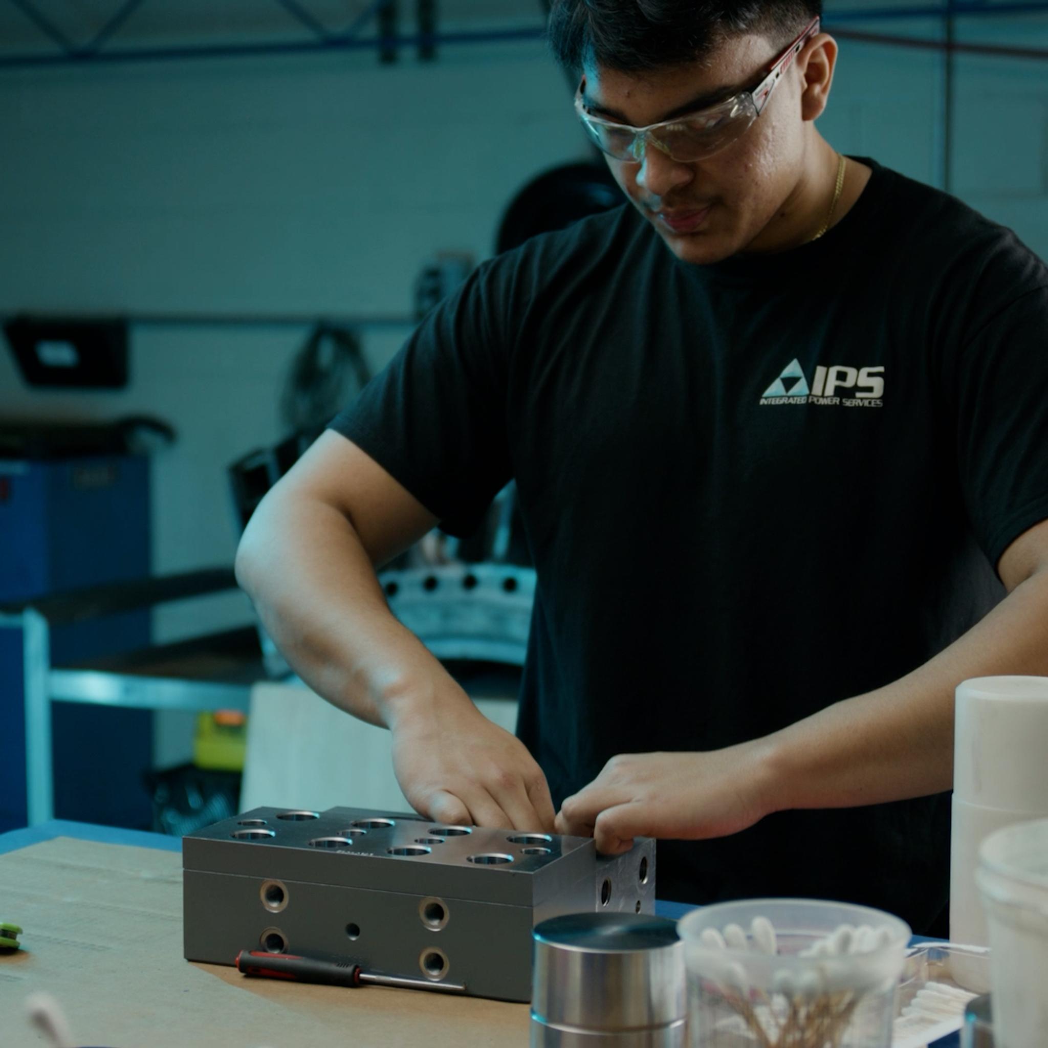 IPS technician wearing safety glasses assembles a metal component at a workbench beside a cordless drill.