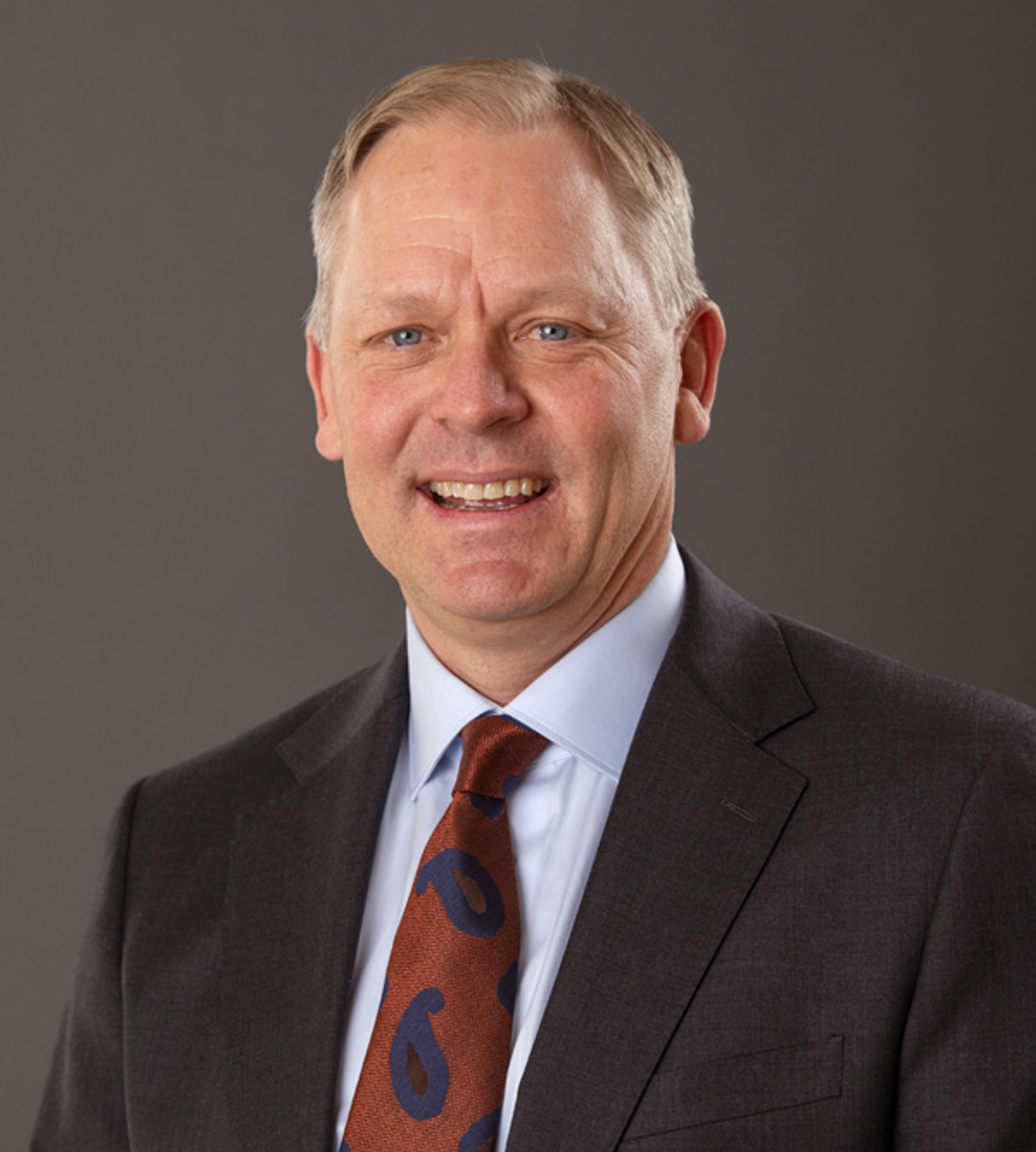 Smiling business executive in a dark suit and patterned tie against a gray background.