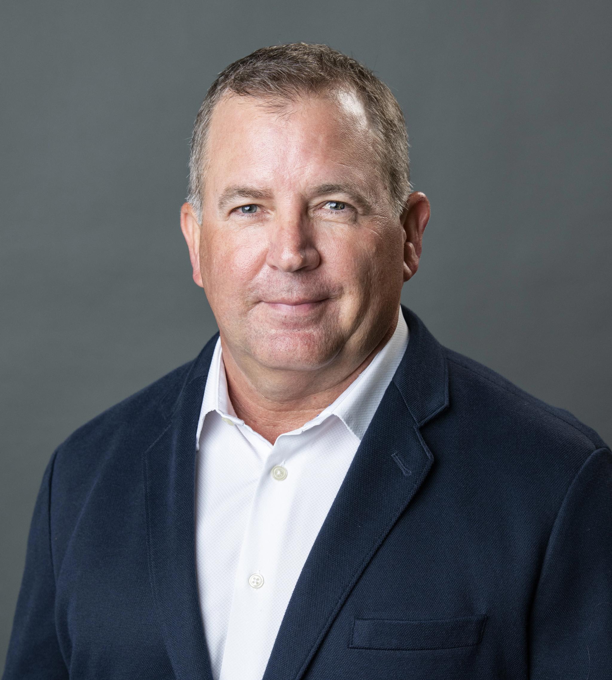 Professional headshot of a man in a navy blazer and white shirt against a gray background.