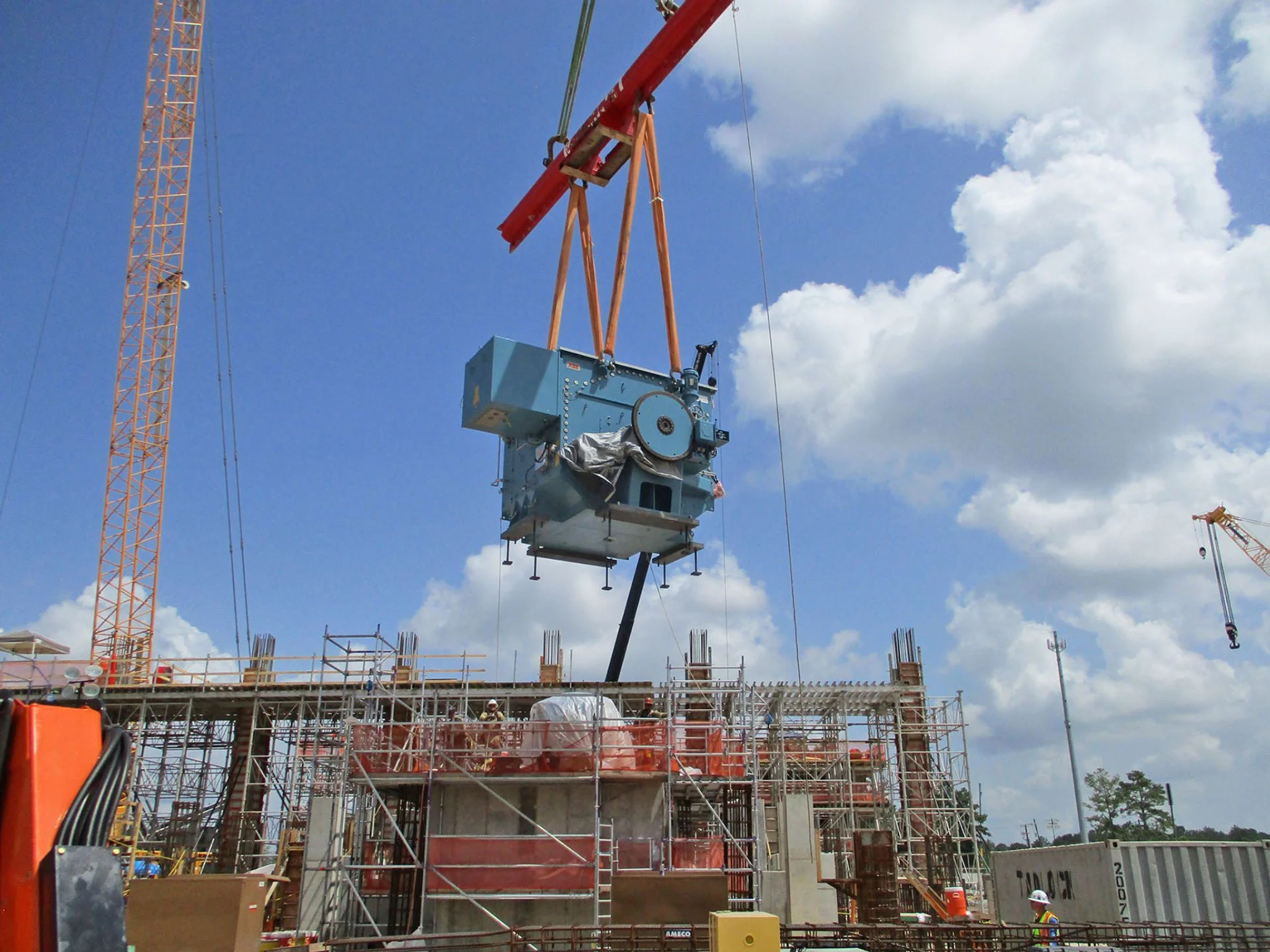 Une grue soulève un transformateur électrique au-dessus d’un chantier industriel.
