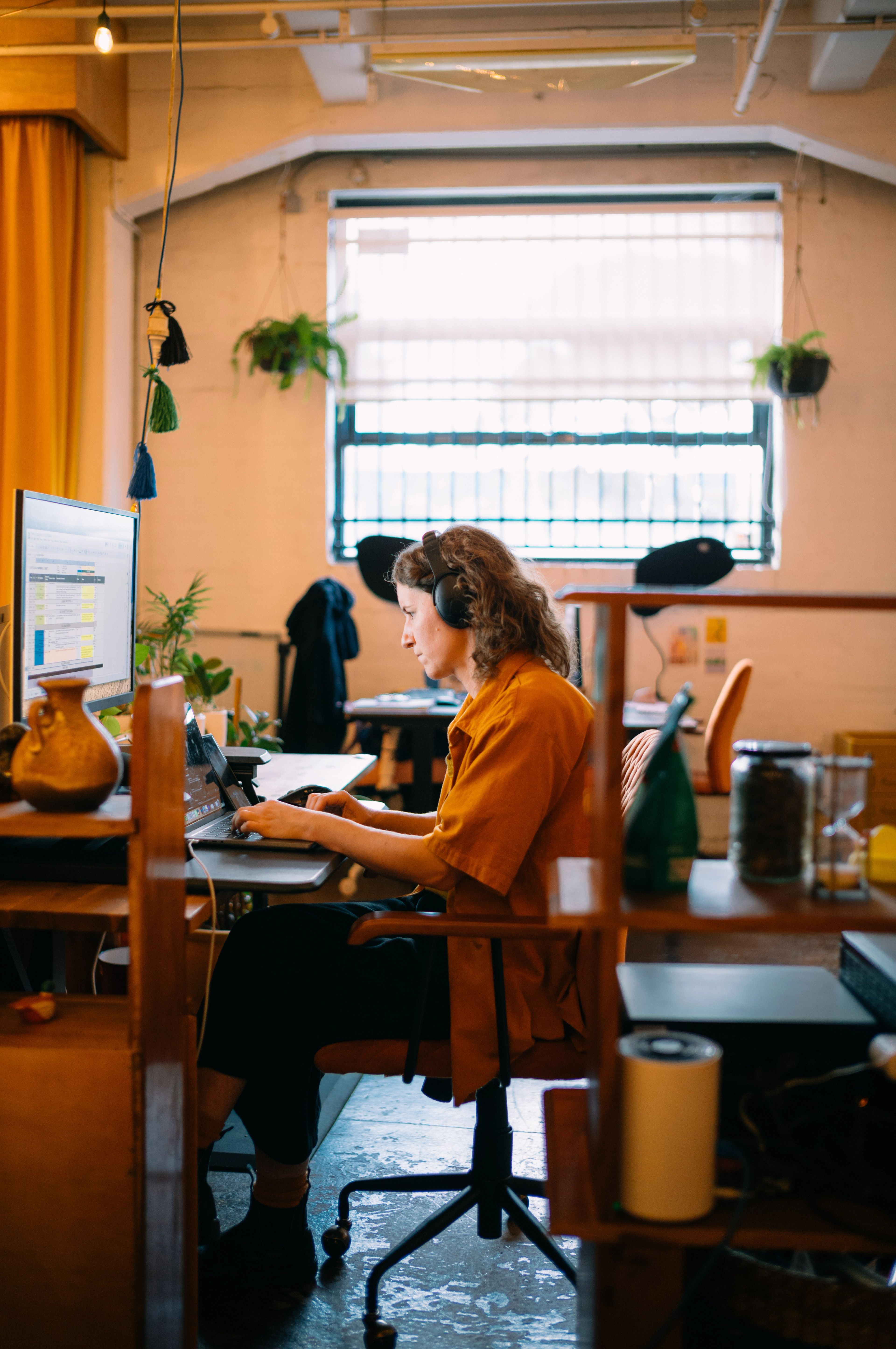 A young woman set up on a wooden desk with his computer, monitor, and knick knacks. Seated at a chair.