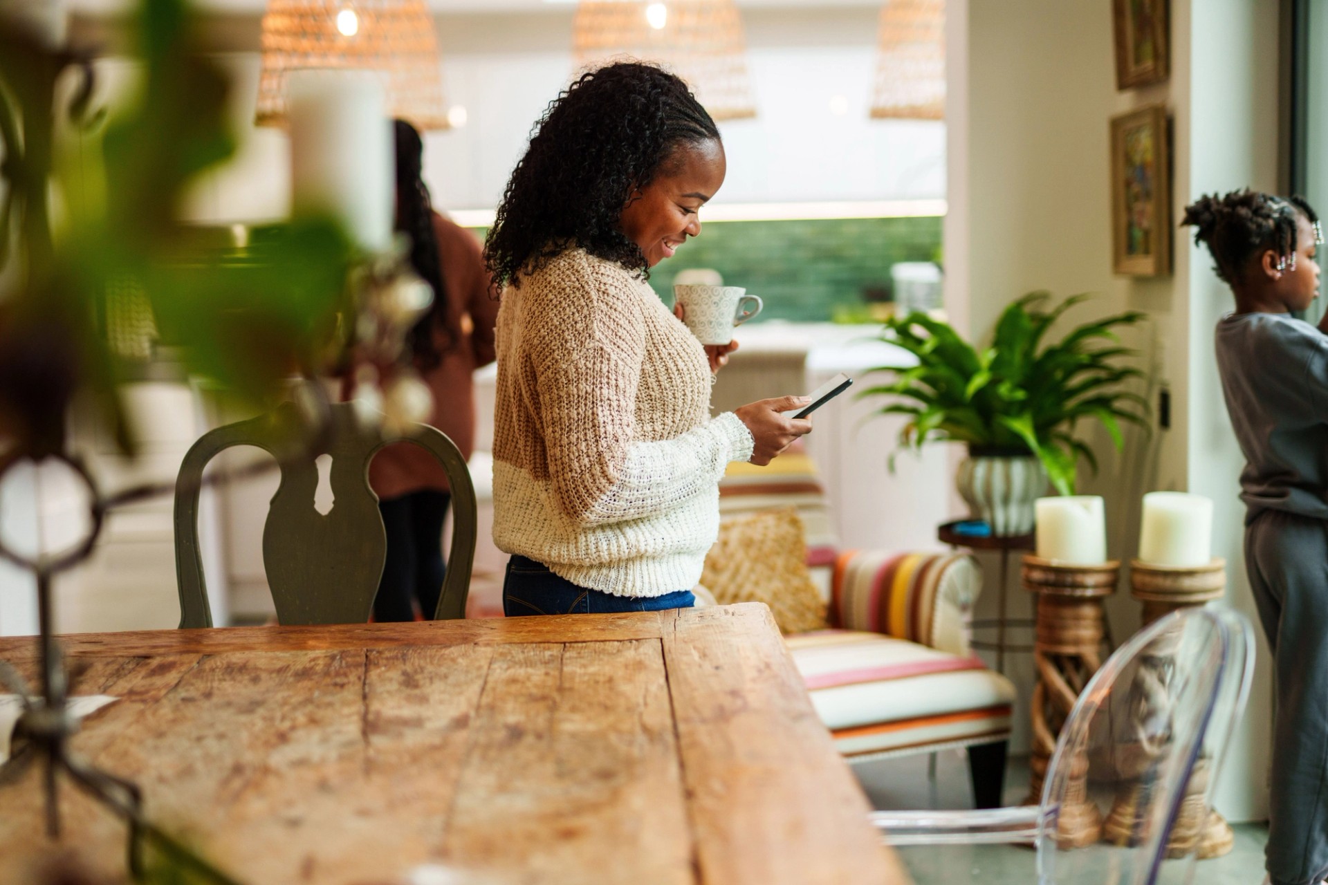 Person using a mobile phone while holding a mug, with family members at home.