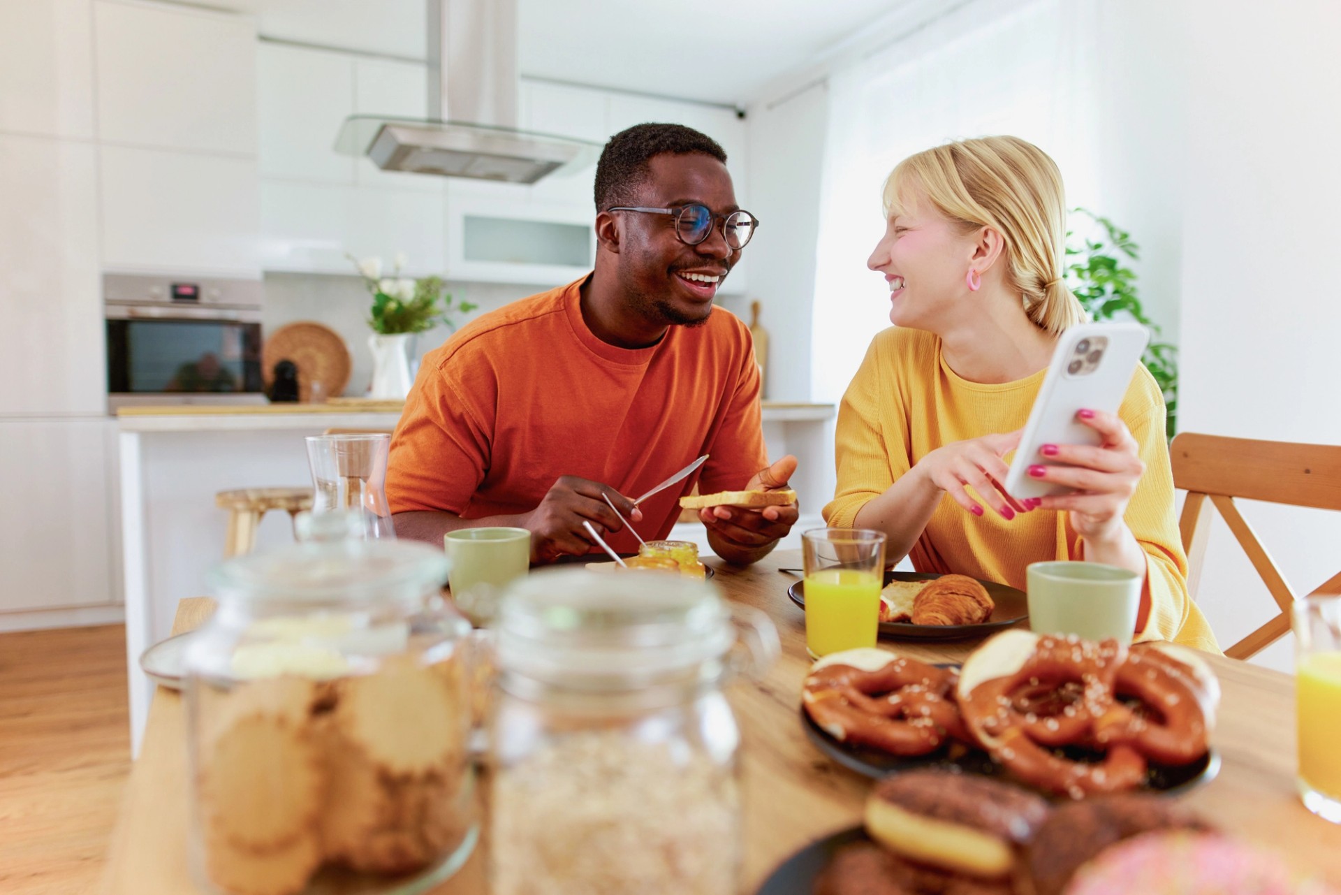 Two people seated at a dining table using a mobile phone during breakfast in a home kitchen.