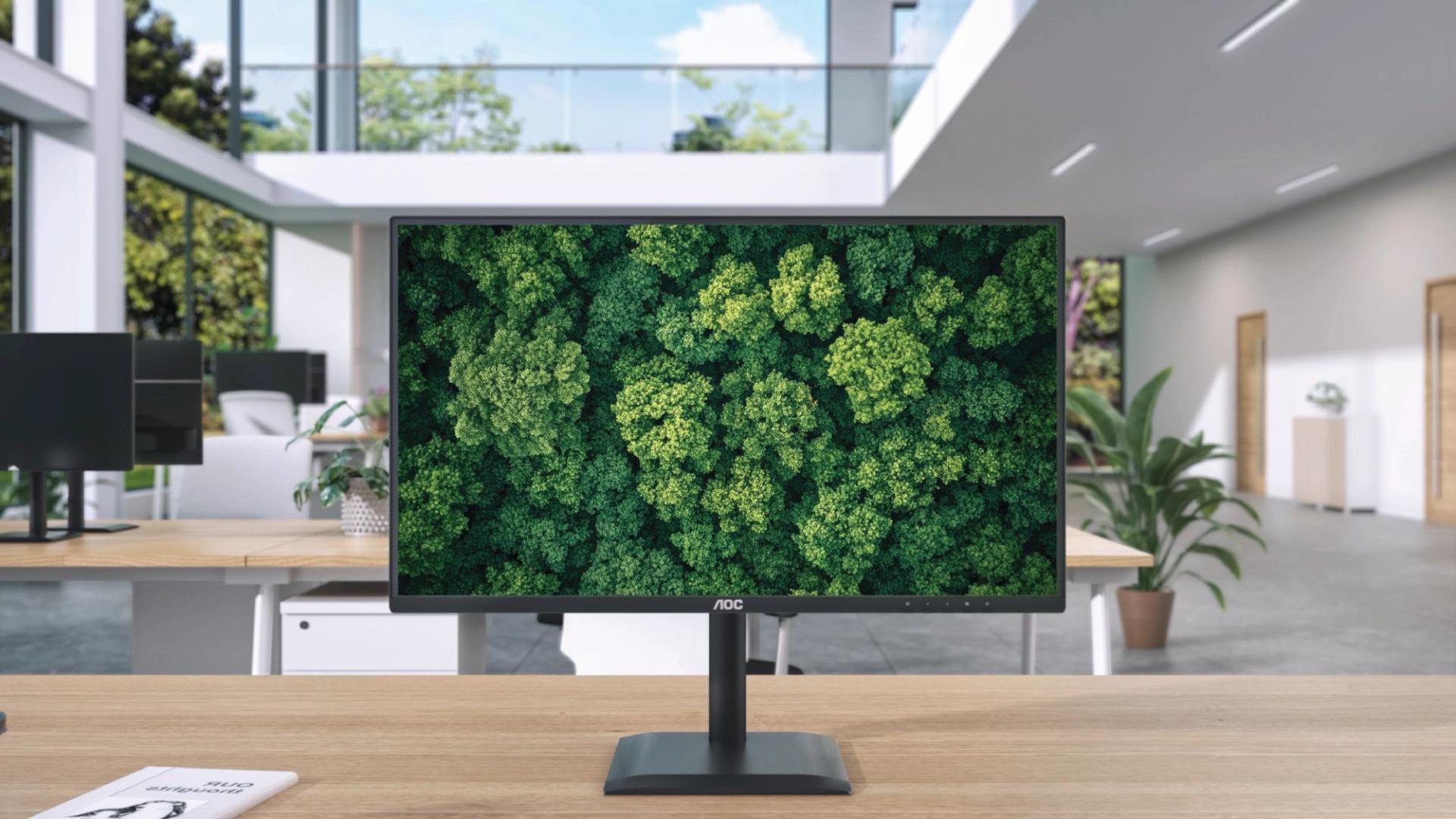 AOC professional monitor displaying a vibrant aerial forest landscape on a desk in a modern open-plan office with large windows, natural light, and minimalist workspace design.