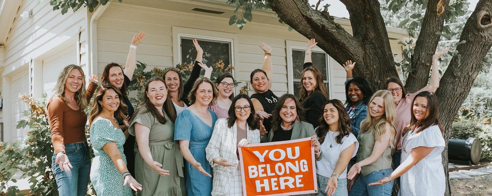 All Moms board members gathered in front of a house holding a 'You Belong Here' sign.