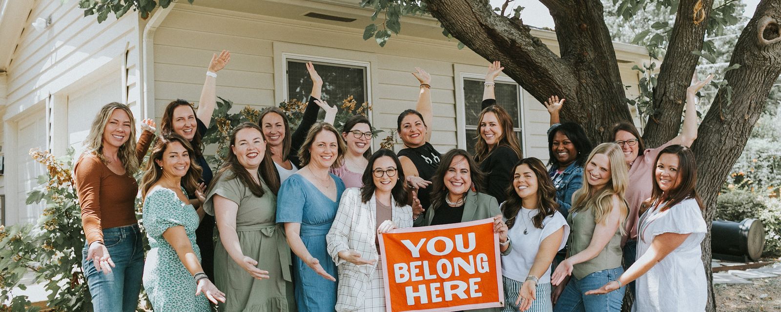 All Moms board members gathered in front of a house holding a 'You Belong Here' sign.