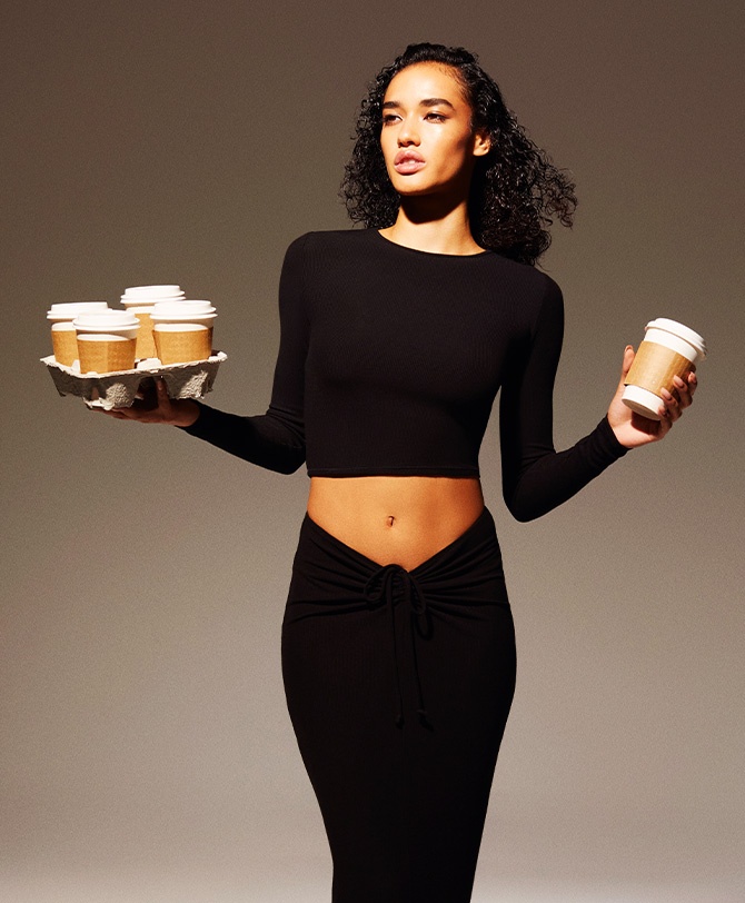 A MODEL POSES WTH COFFEE CUPS, WEARING SKIMS SOFT LOUNGE
