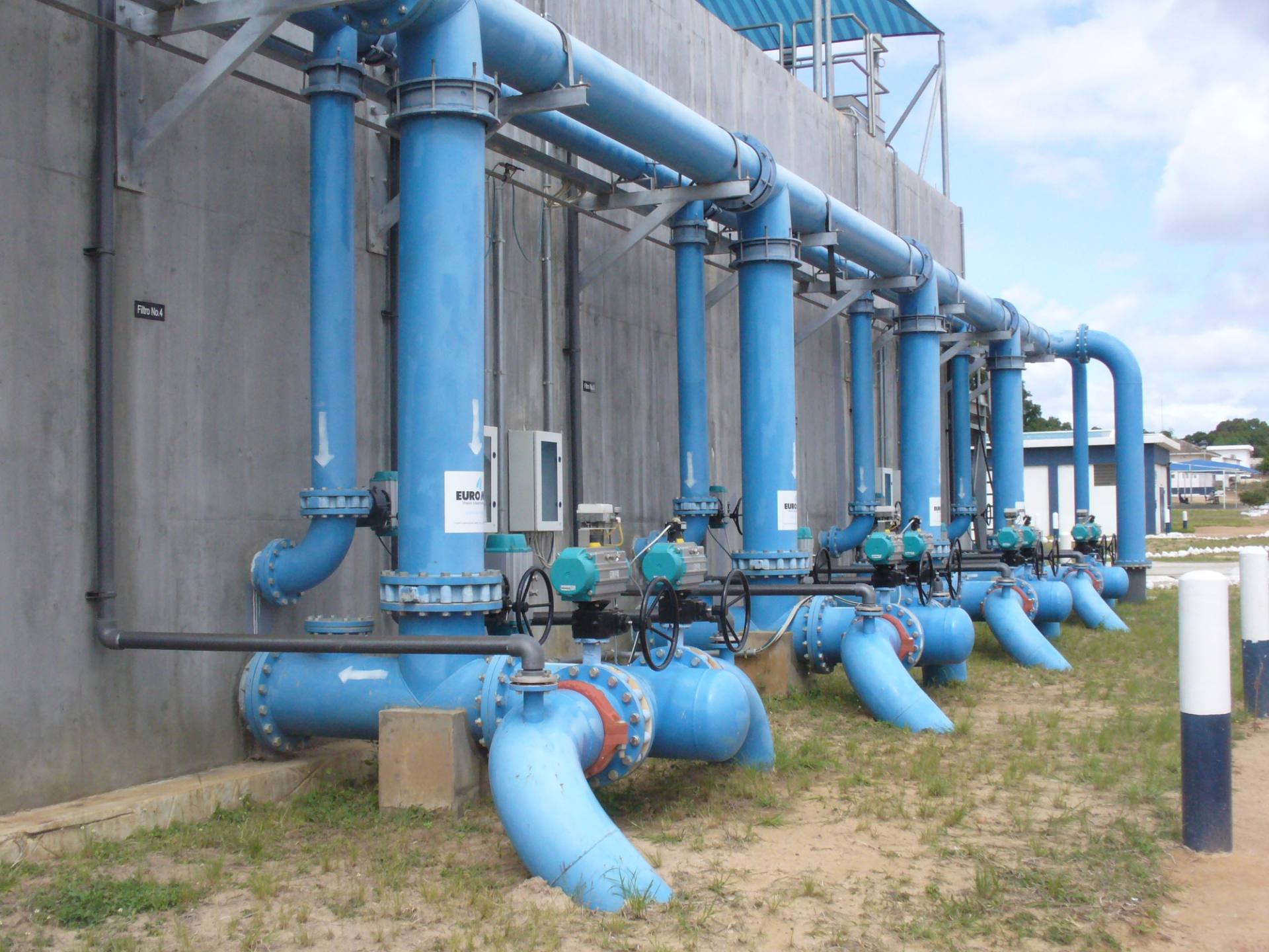 A row of blue steel pipes and valves at a water treatment plant, under a bright sky.