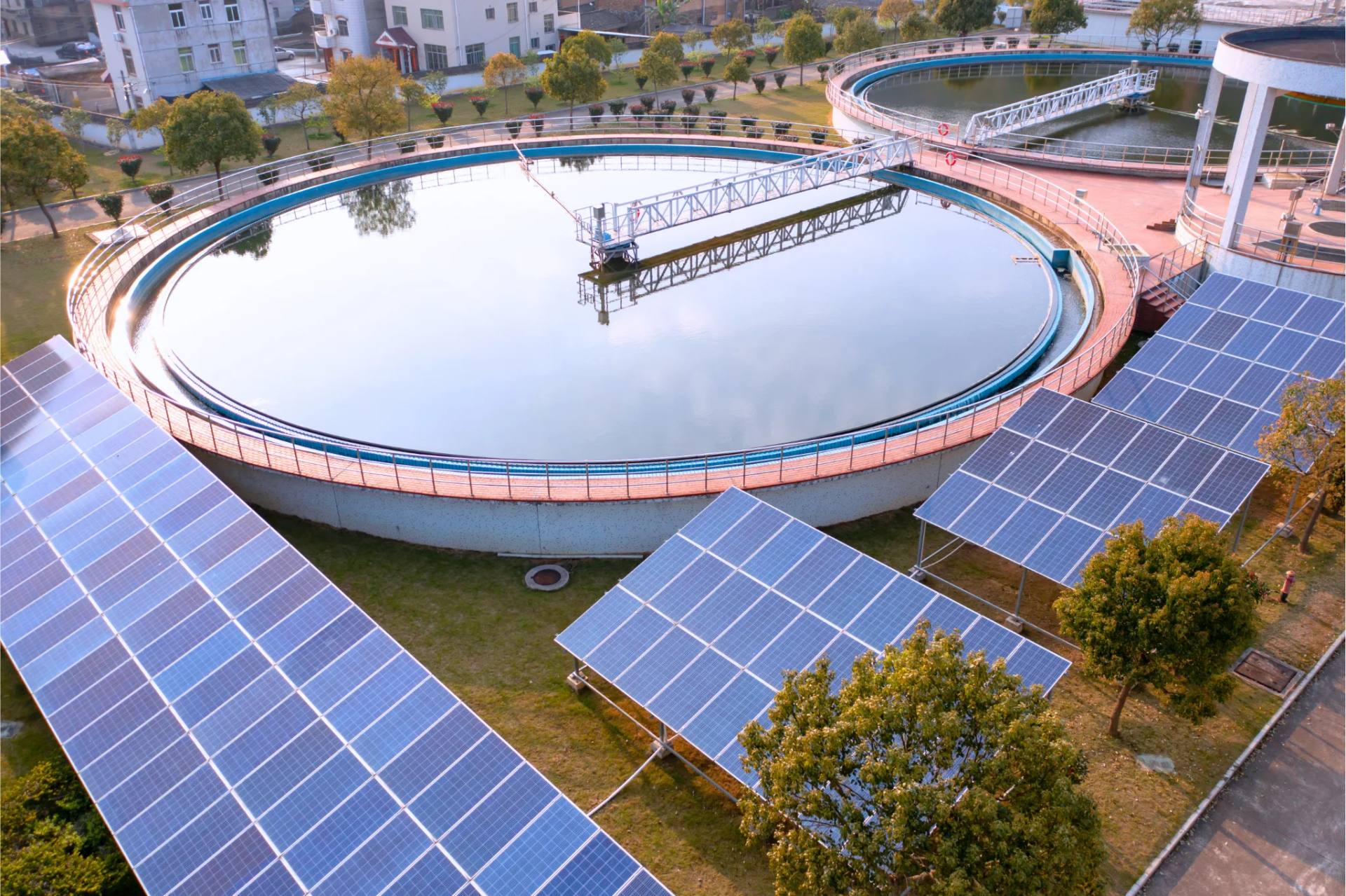 Aerial view of a water treatment plant: a circular sedimentation tank surrounded by rows of solar panels.