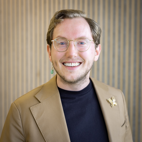 Portrait of Mr Sietse Blom in a beige jacket over a dark top, against a wooden slatted wall.