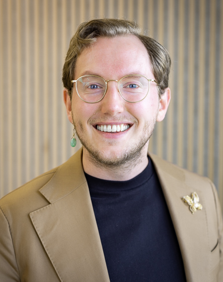 Portrait of Mr Sietse Blom in a beige jacket over a dark top, against a wooden slatted wall.