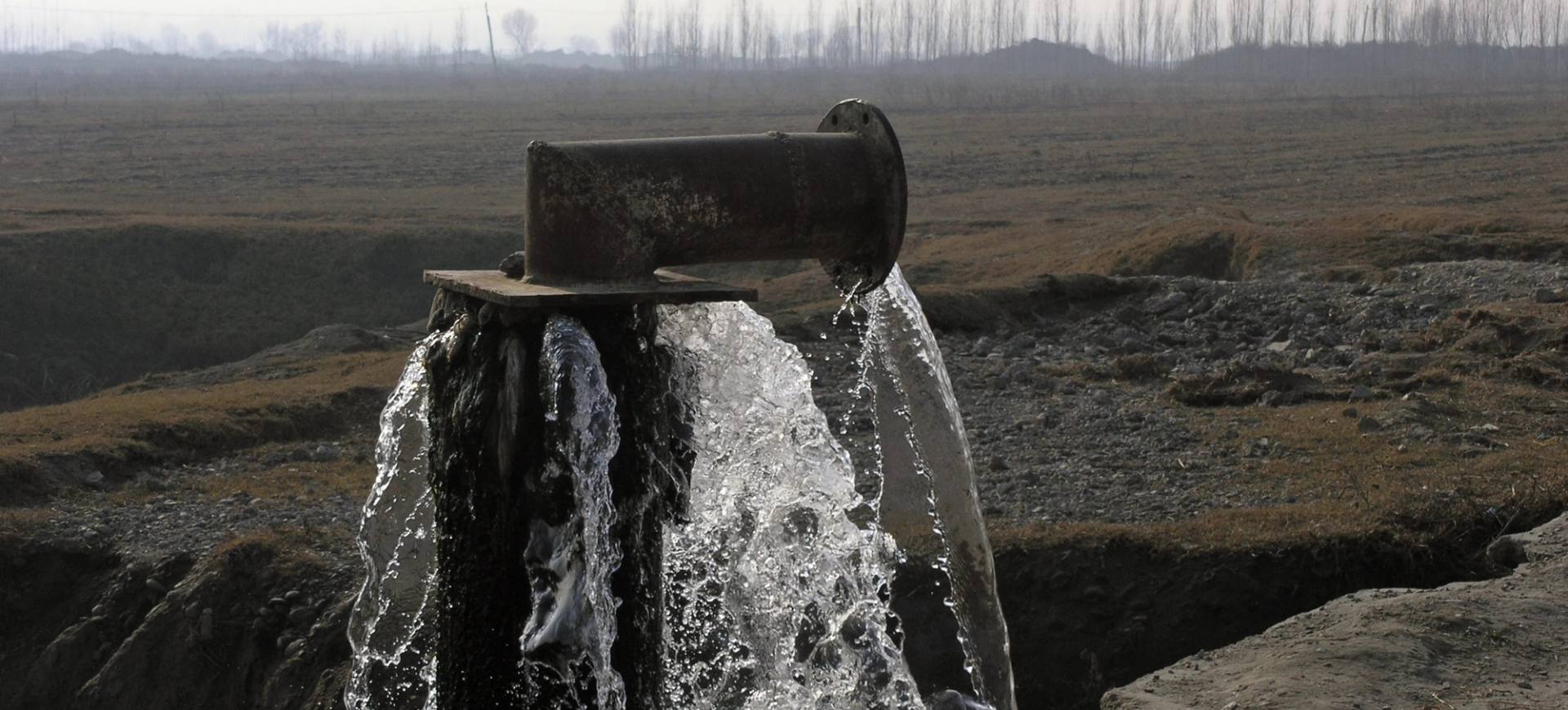 Water gushing from a steel pipe into an irrigation channel in an open landscape.