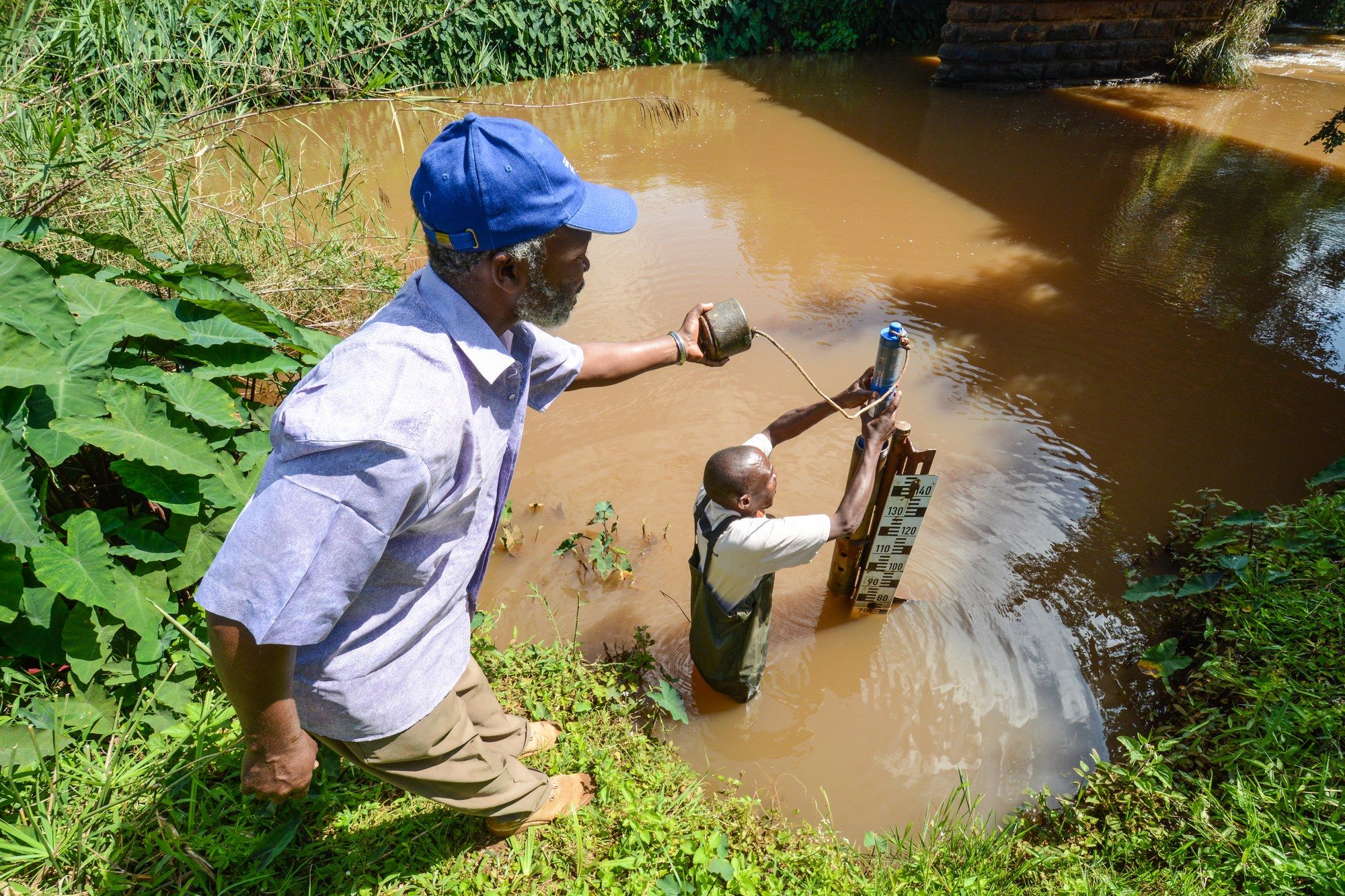 A man and a child collecting water from a pipe at a muddy pond