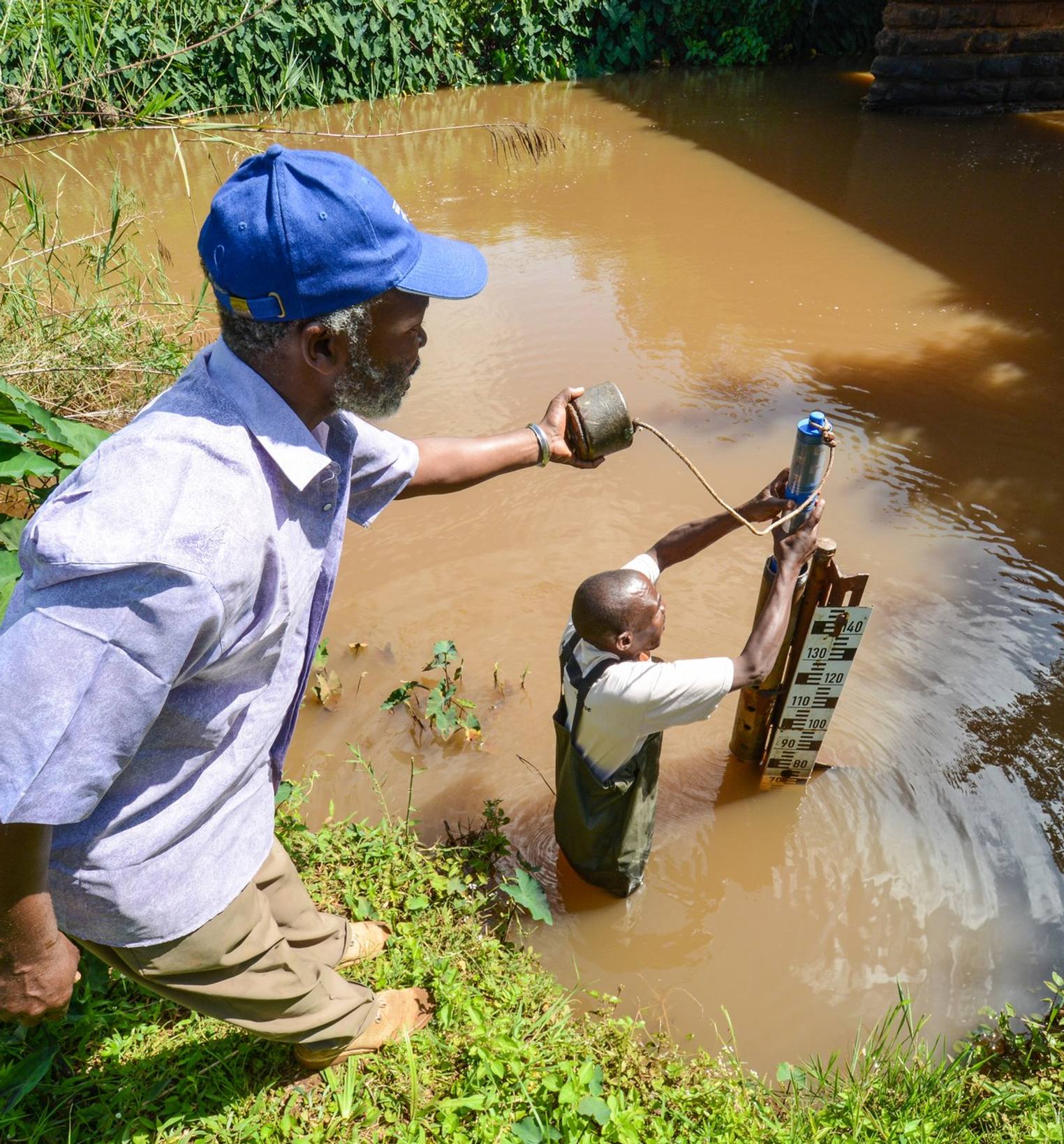 A man and a child collecting water from a pipe at a muddy pond