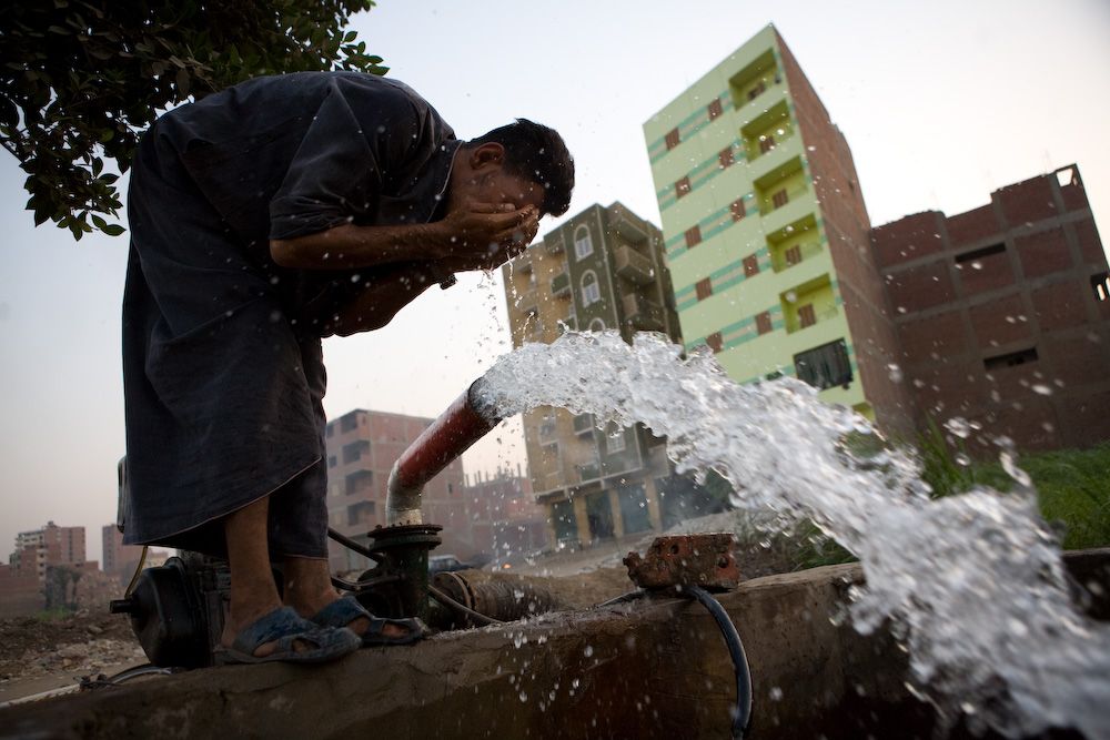 A man washing his face in water gushing from a standpipe in a city street.