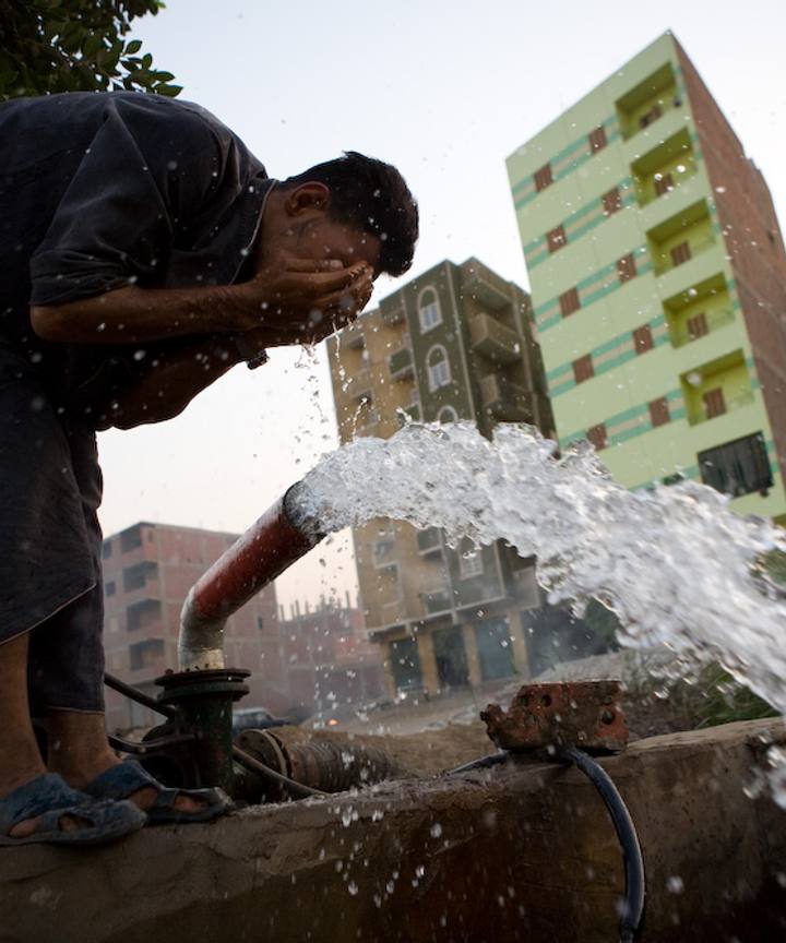 A man washing his face in water gushing from a standpipe in a city street.