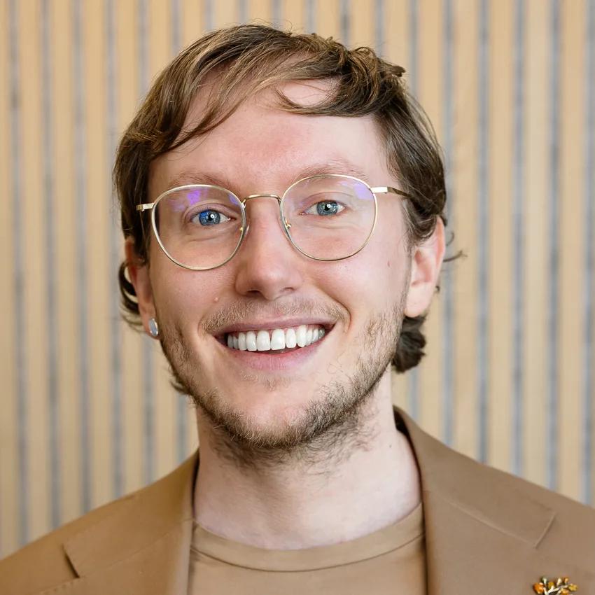 Portrait of Mr Sietse Blom in a beige jacket over a dark top, against a wooden slatted wall.
