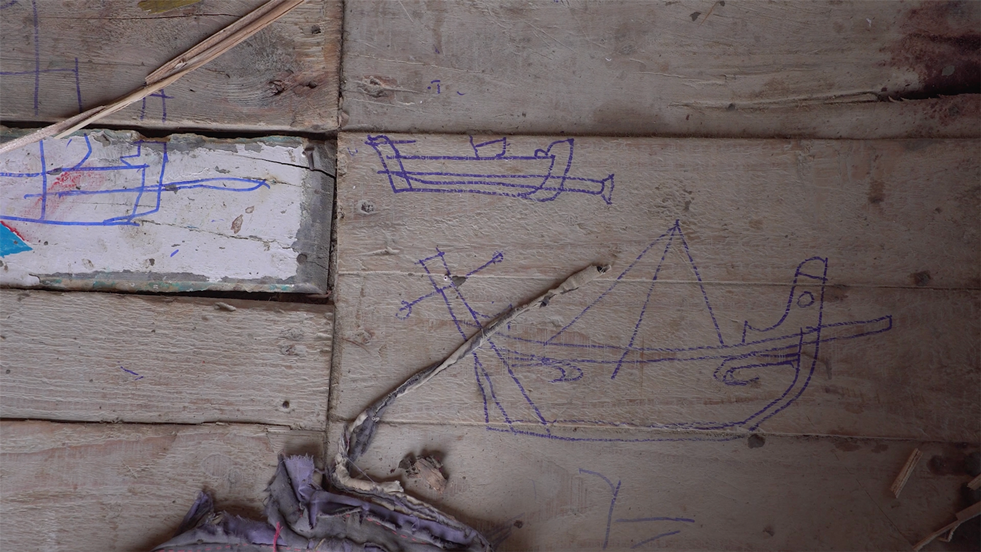 3 boats drawn in blue pen on a wooden floor, with ripped cloth in the foreground.