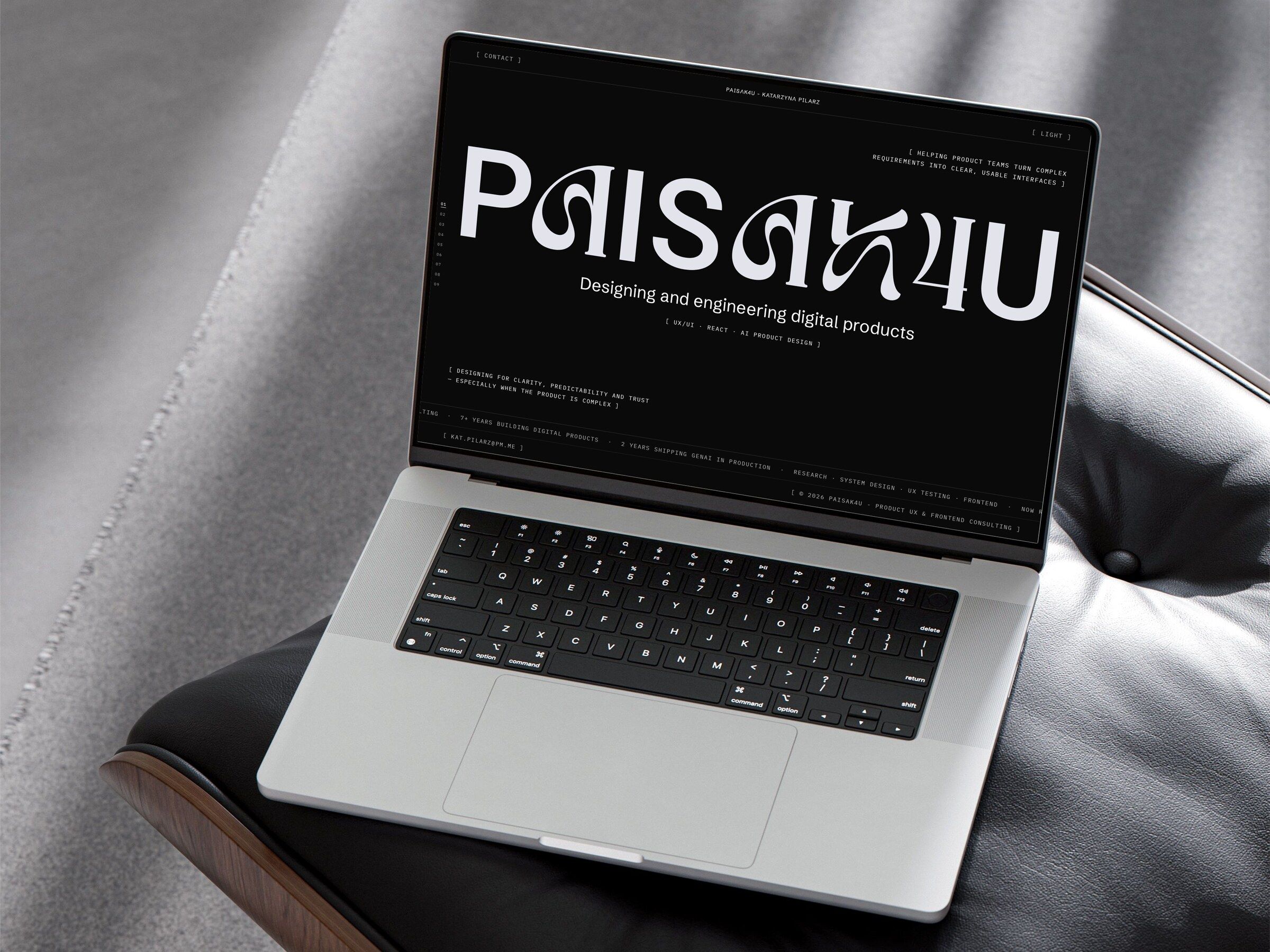 The PAISAK4U home page in the dark theme on a laptop resting on a black leather lounge chair